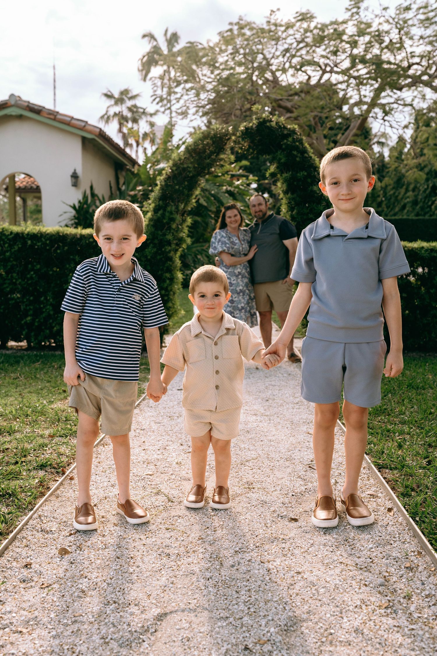 Three young brothers hold hands on a garden path while their parents stand together in the background.  Family photos at Bradley Park
