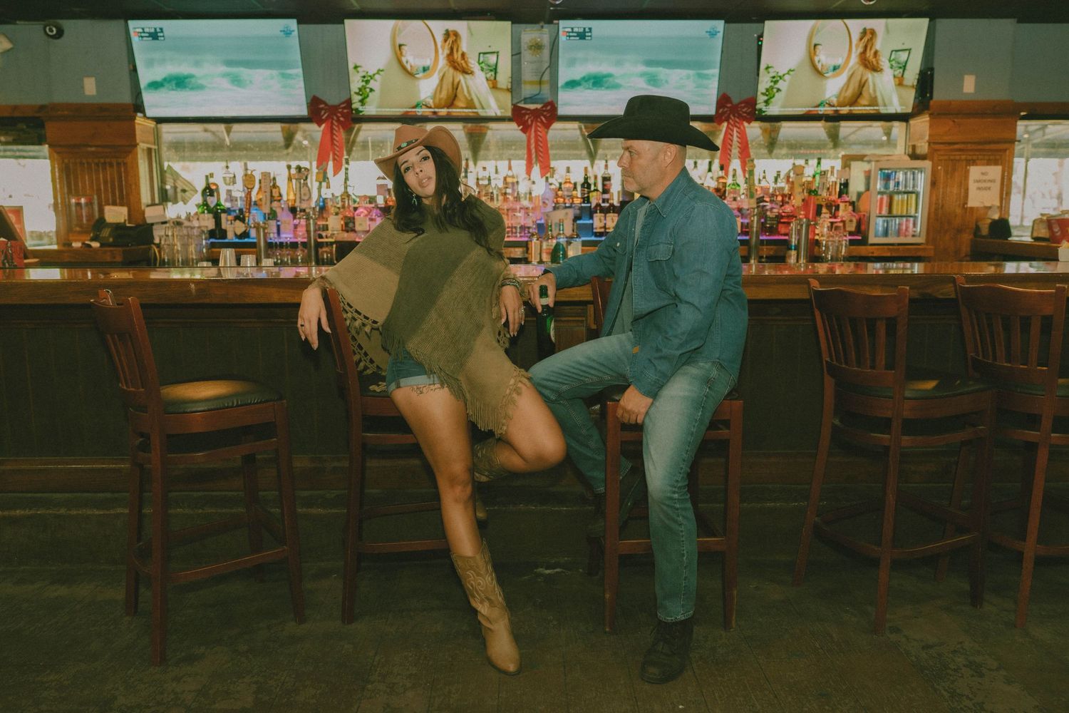 A man in a cowboy hat and woman in a poncho pose at a rustic bar with colorful bottles and TVs.
