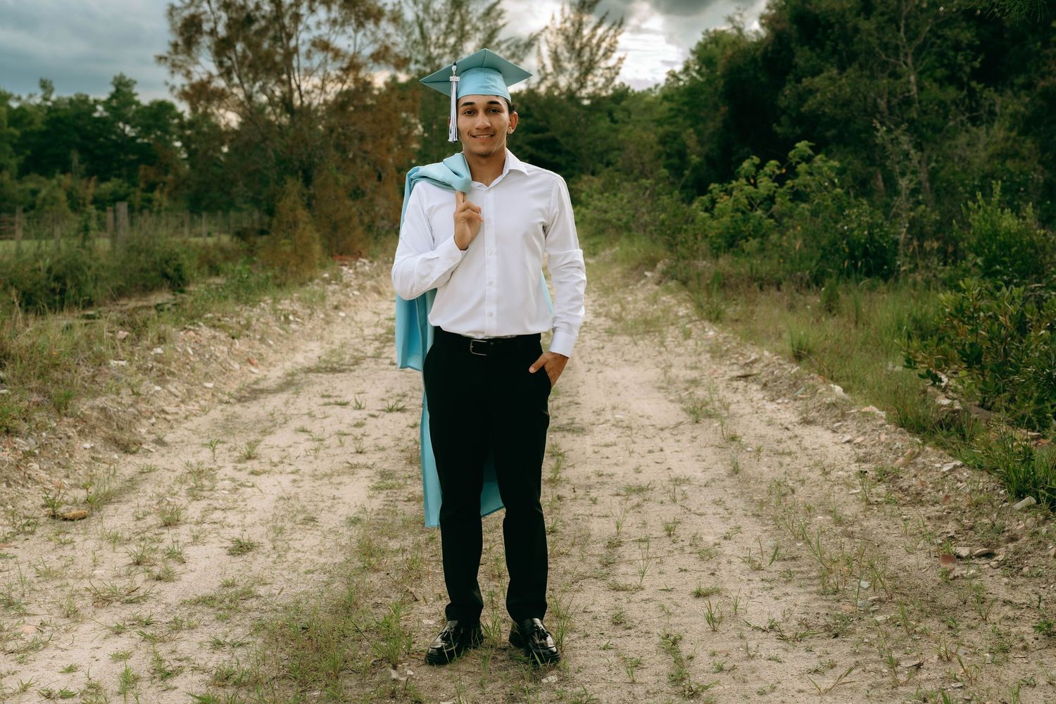 Graduate in light blue cap and gown poses on a dirt path surrounded by lush greenery. Senior Photos at Colt Farms in Boynton Beach, FL