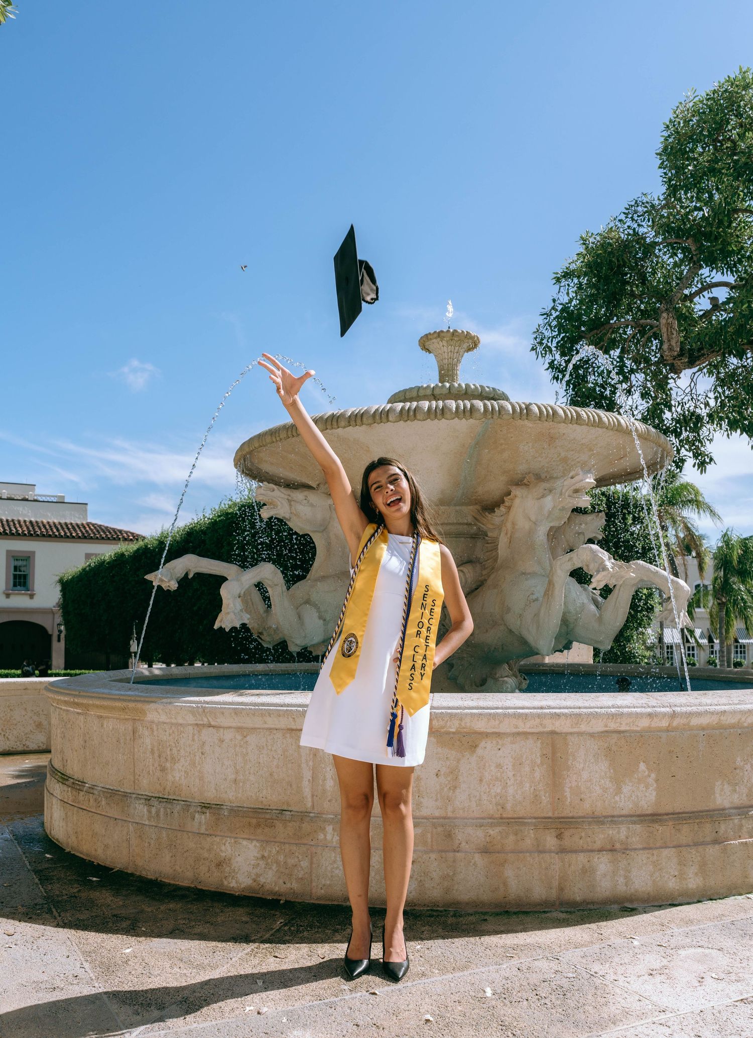 Smiling graduate tosses cap in air by decorative stone fountain in bright blue sky on Worth Avenue Palm beach.