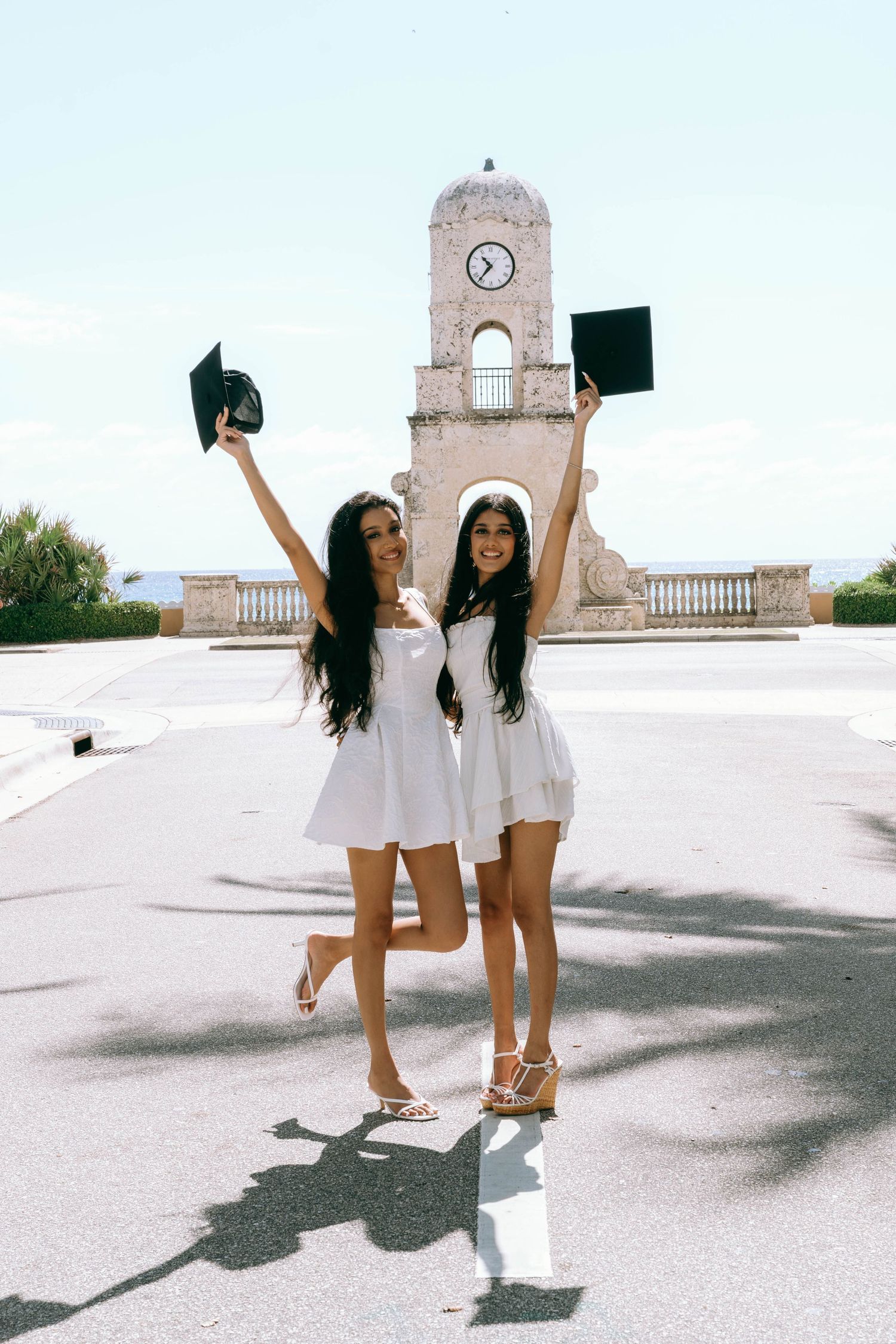 Two young women in white dresses celebrate graduation by raising their caps near a clock tower on Worth Avenue Palm beach.