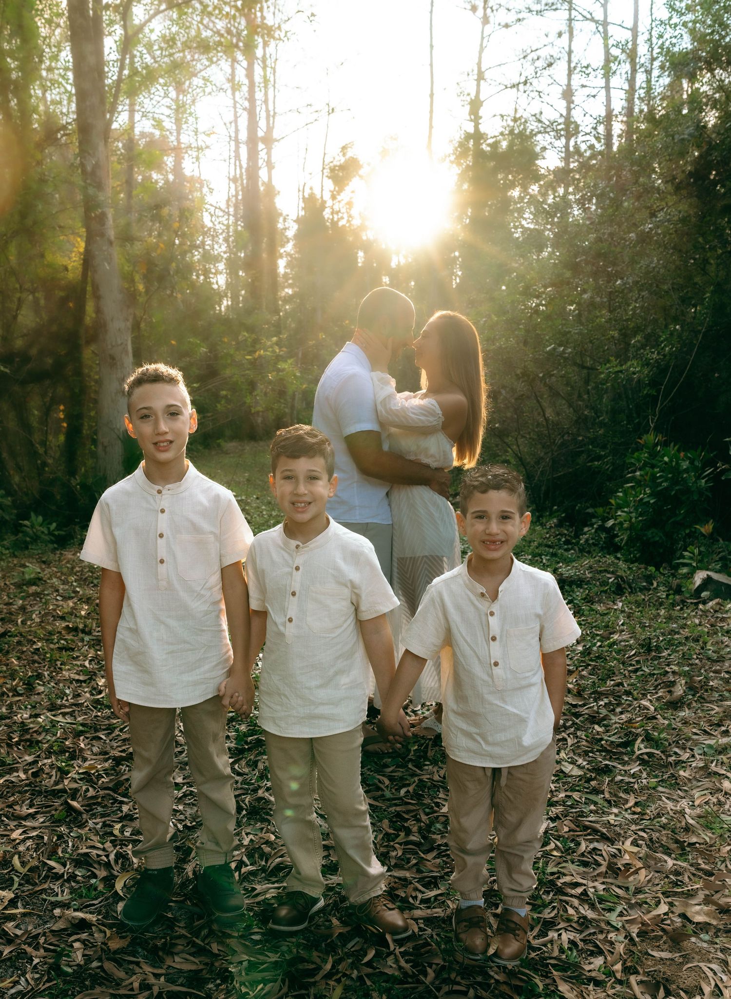 Three children in white shirts and khaki pants stand together in a sunlit wooded area during golden hour.