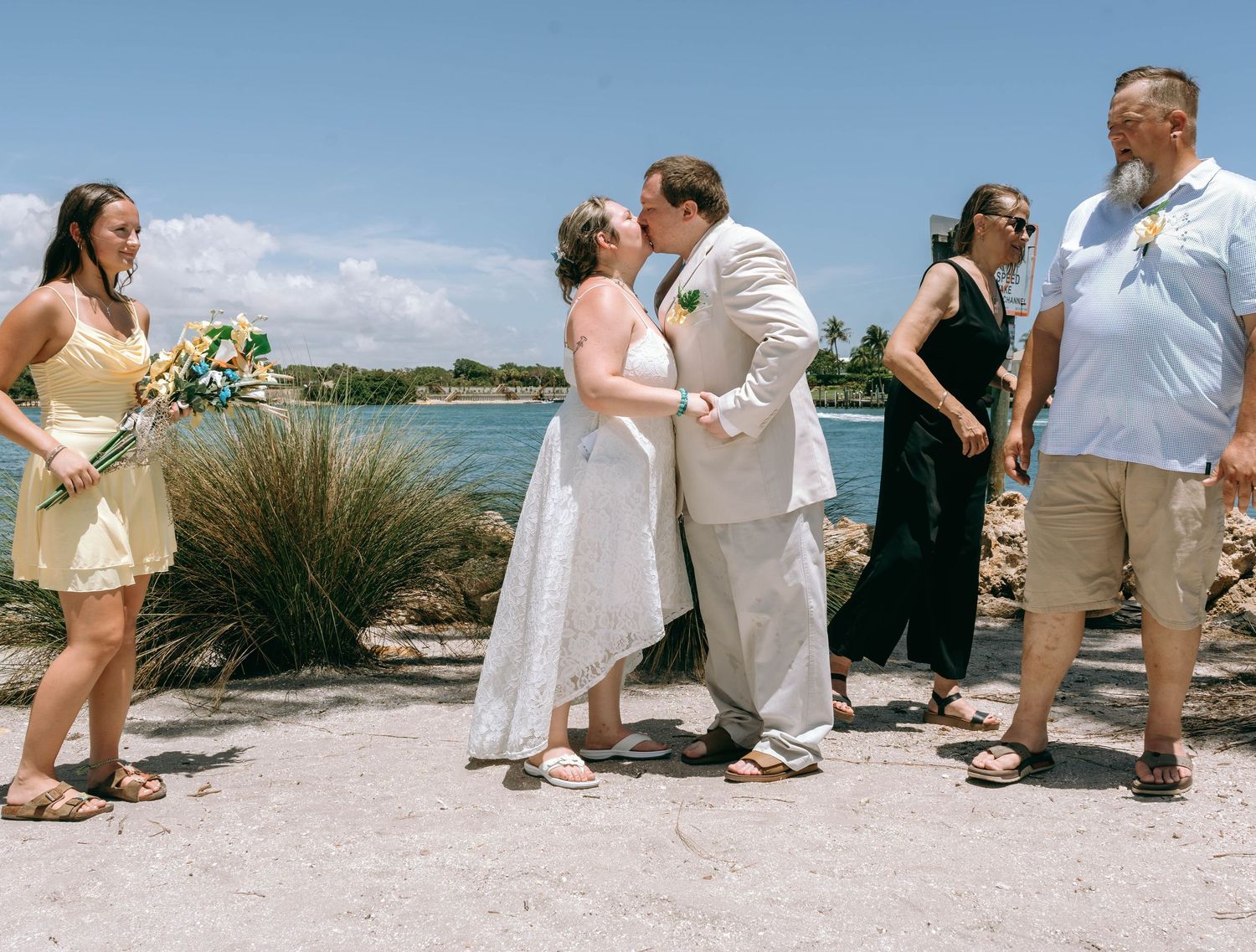 Bride and groom share a kiss at outdoor beach wedding ceremony with guests watching near tropical waterfront.