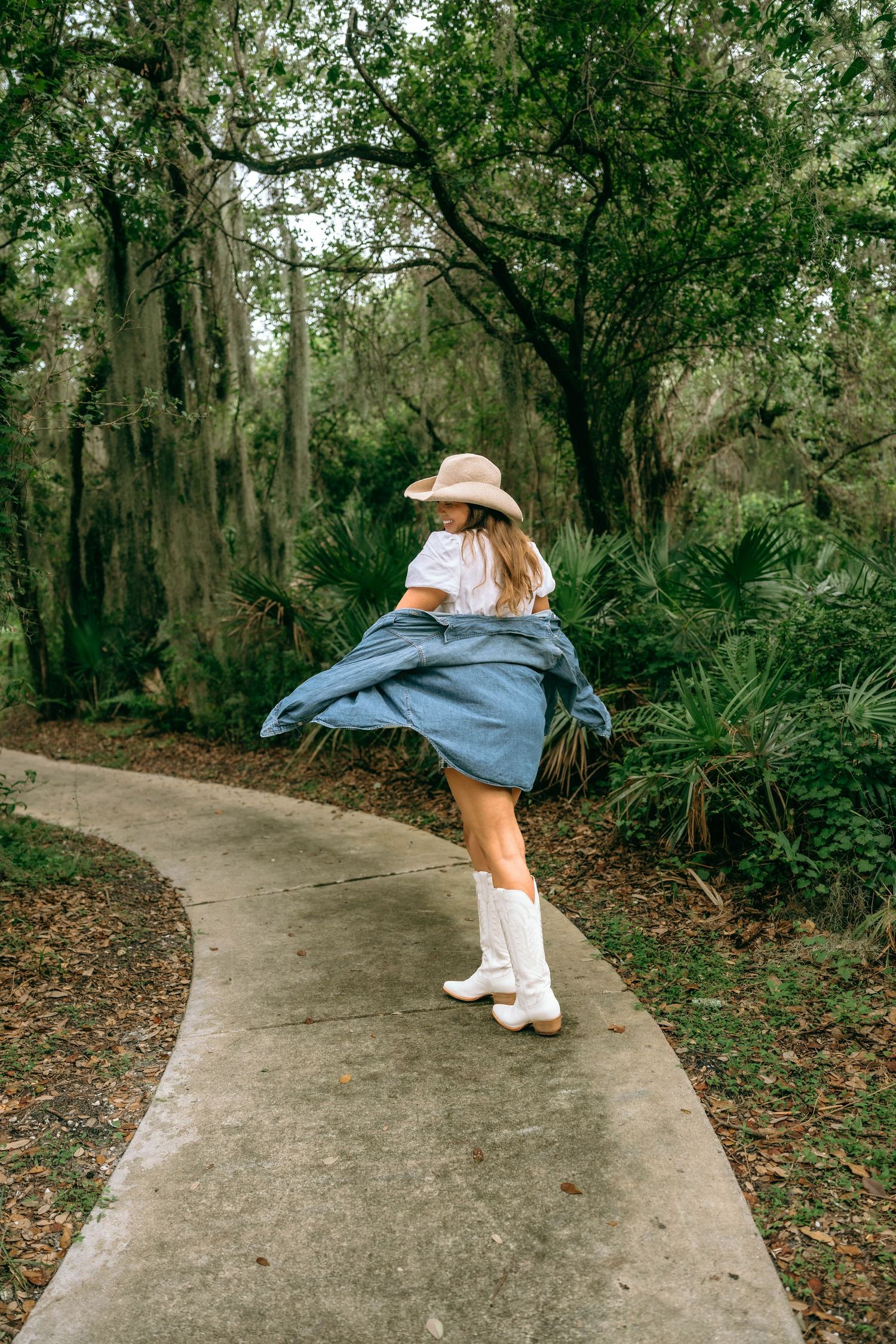 Woman in white top, denim skirt, cowboy hat, and white boots twirling on a path in a lush green forest.