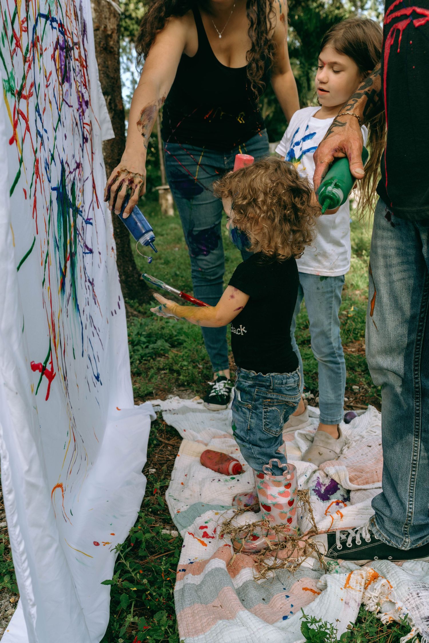 Children and adults painting on a white canvas outdoors, with colorful paint splatters on fabric and hands.