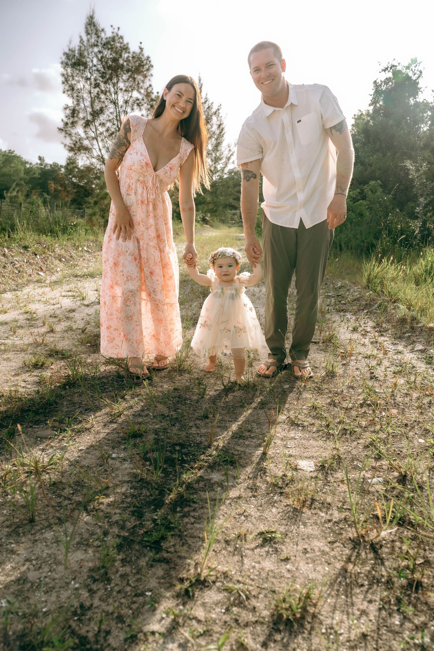 Happy young family with toddler daughter holding hands on a sunny nature trail, wearing summer dresses and casual attire.