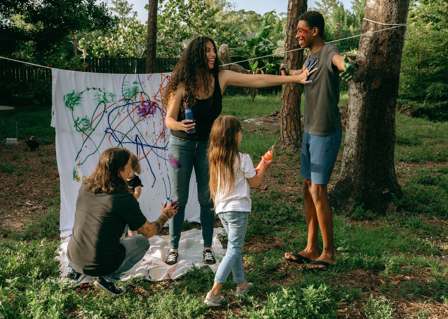 Family painting colorful artwork on a white canvas hung outdoors between trees in a lush green backyard.