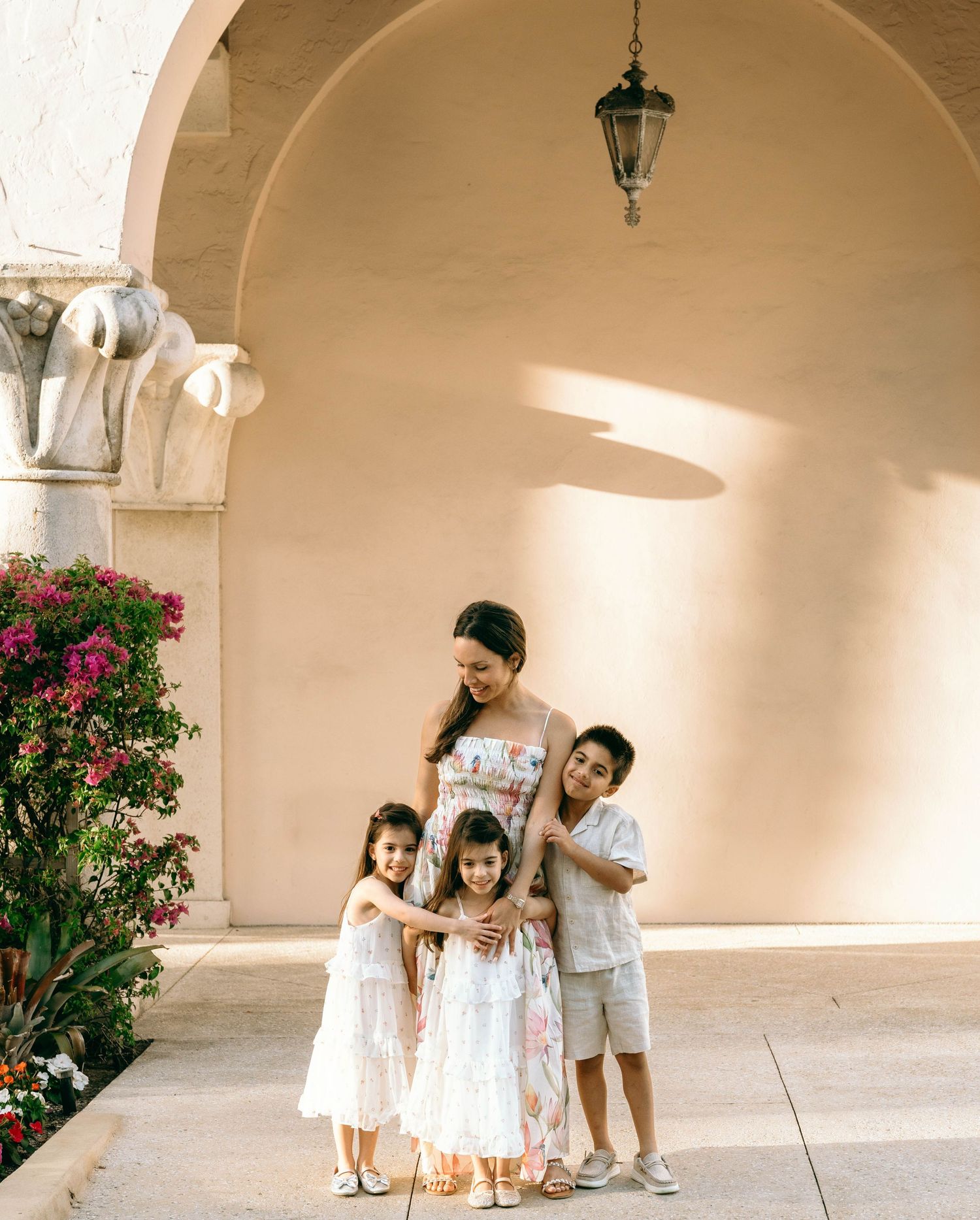A mother in a floral maxi dress embraces her three children under an arched colonnade with blooming flowers. Worth Avenue, Palm Beach, FL