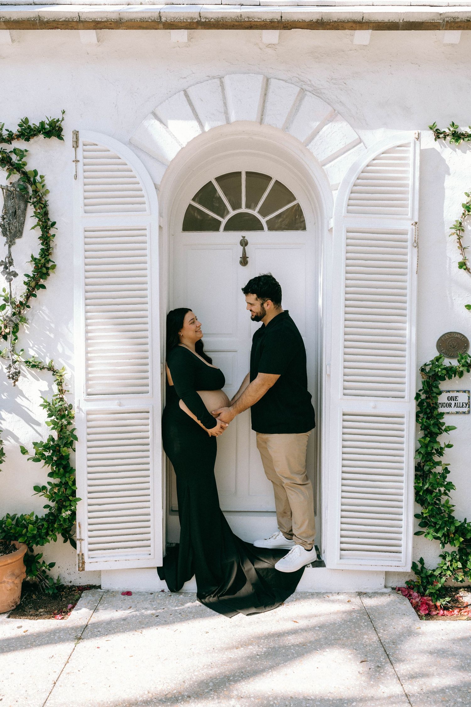 Pregnant couple holding hands in a black dress and casual outfit standing in a white arched doorway with greenery. Worth Avenue, Palm Beach,