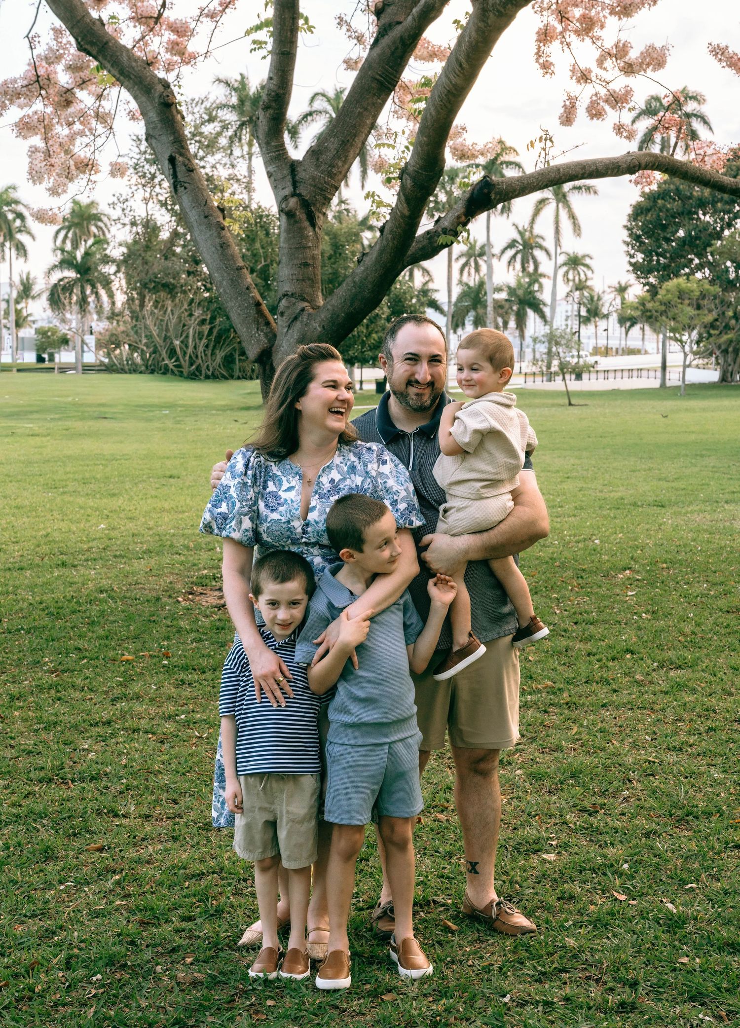 Family of five smiling outdoors under a blooming tree on green grass.  Family photos at Bradley Park Palm Beach, FL