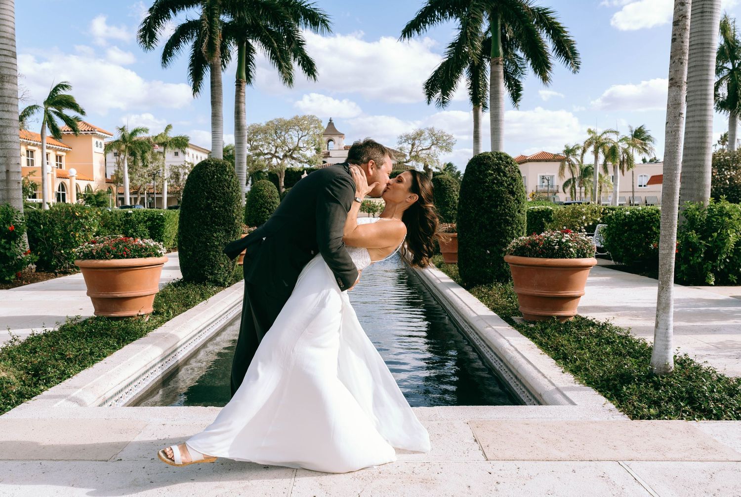 Bride and groom sharing a romantic kiss beside a reflecting pool surrounded by palm trees at an elegant outdoor venue.
