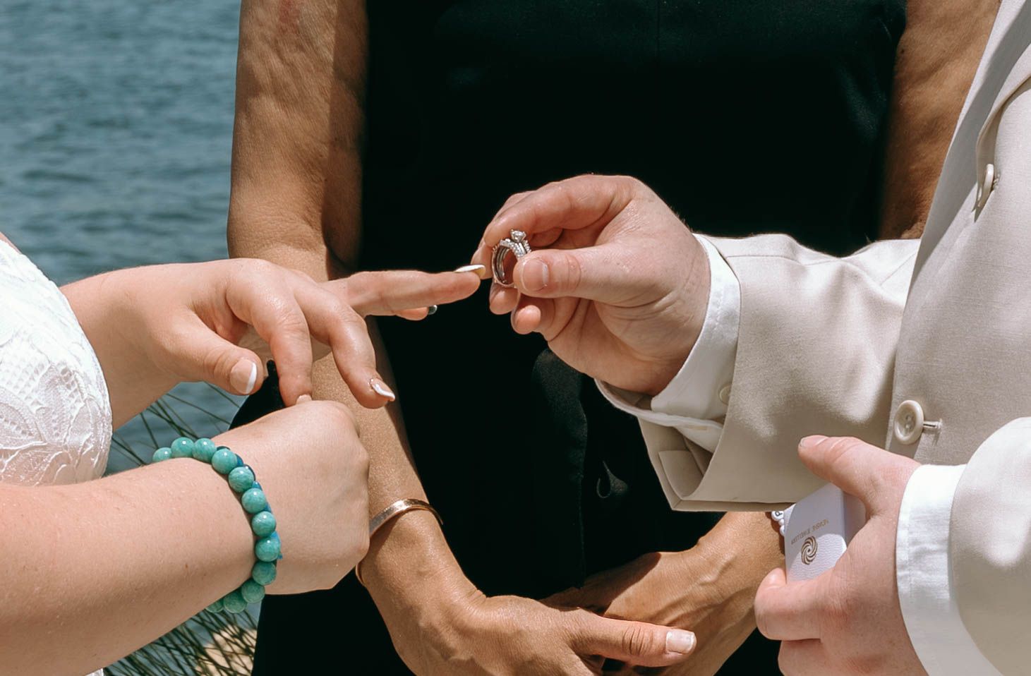 Close-up of wedding ring exchange ceremony with guests wearing green bracelet near water.