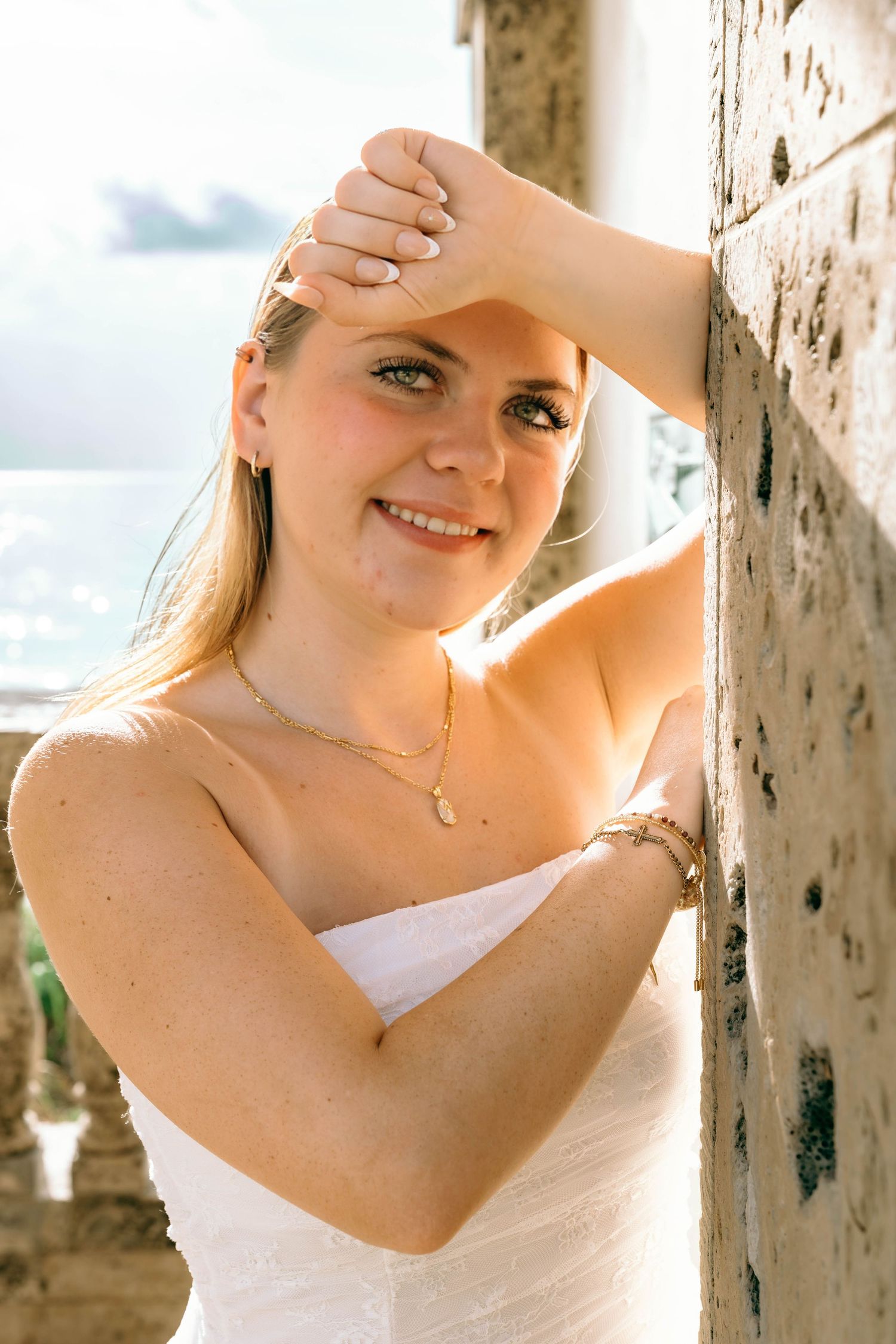 Smiling blonde woman in white strapless dress leaning against stone pillar, wearing gold necklace and bracelet.