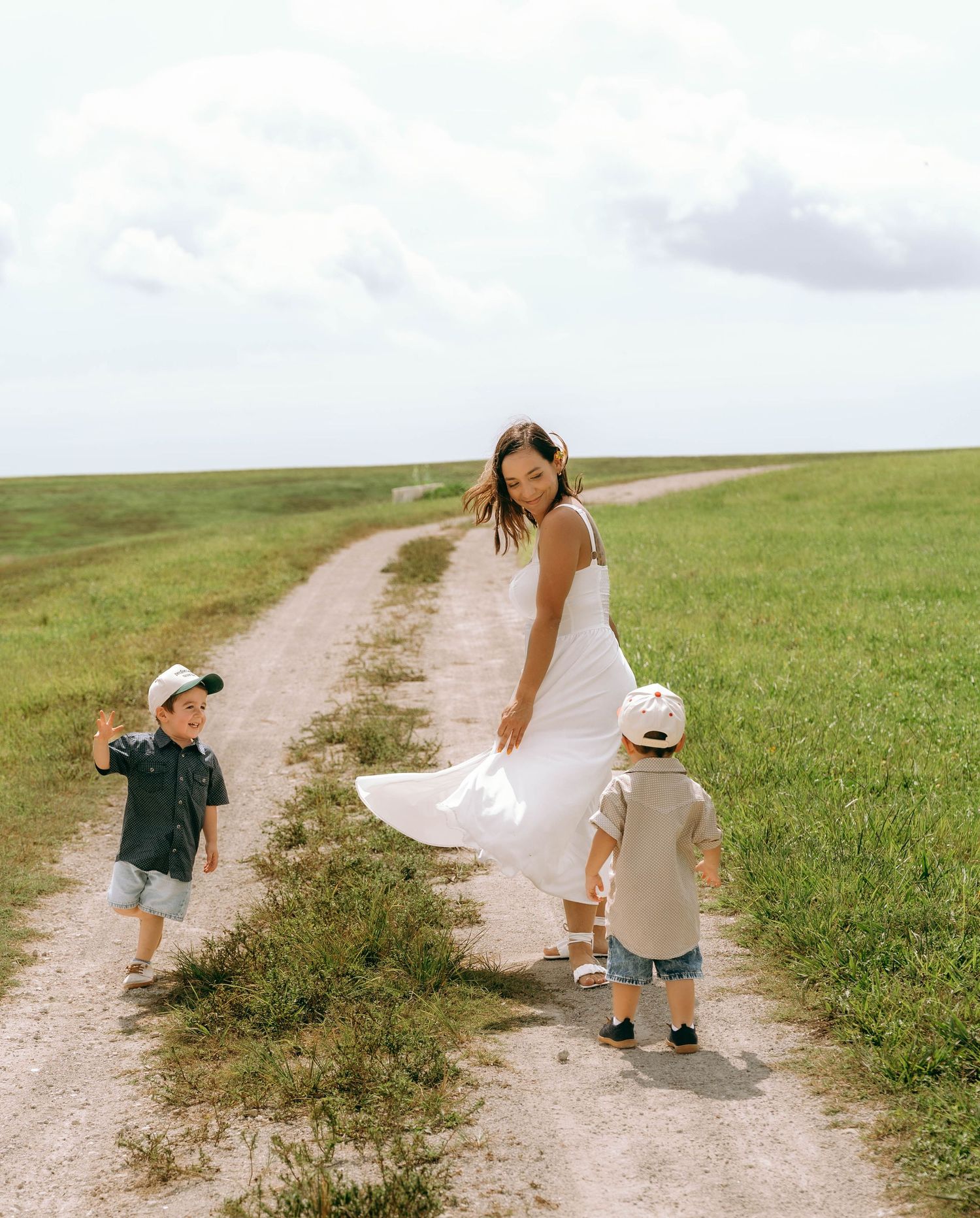 A woman in a white dress walks a dirt path with two young children in a green field.