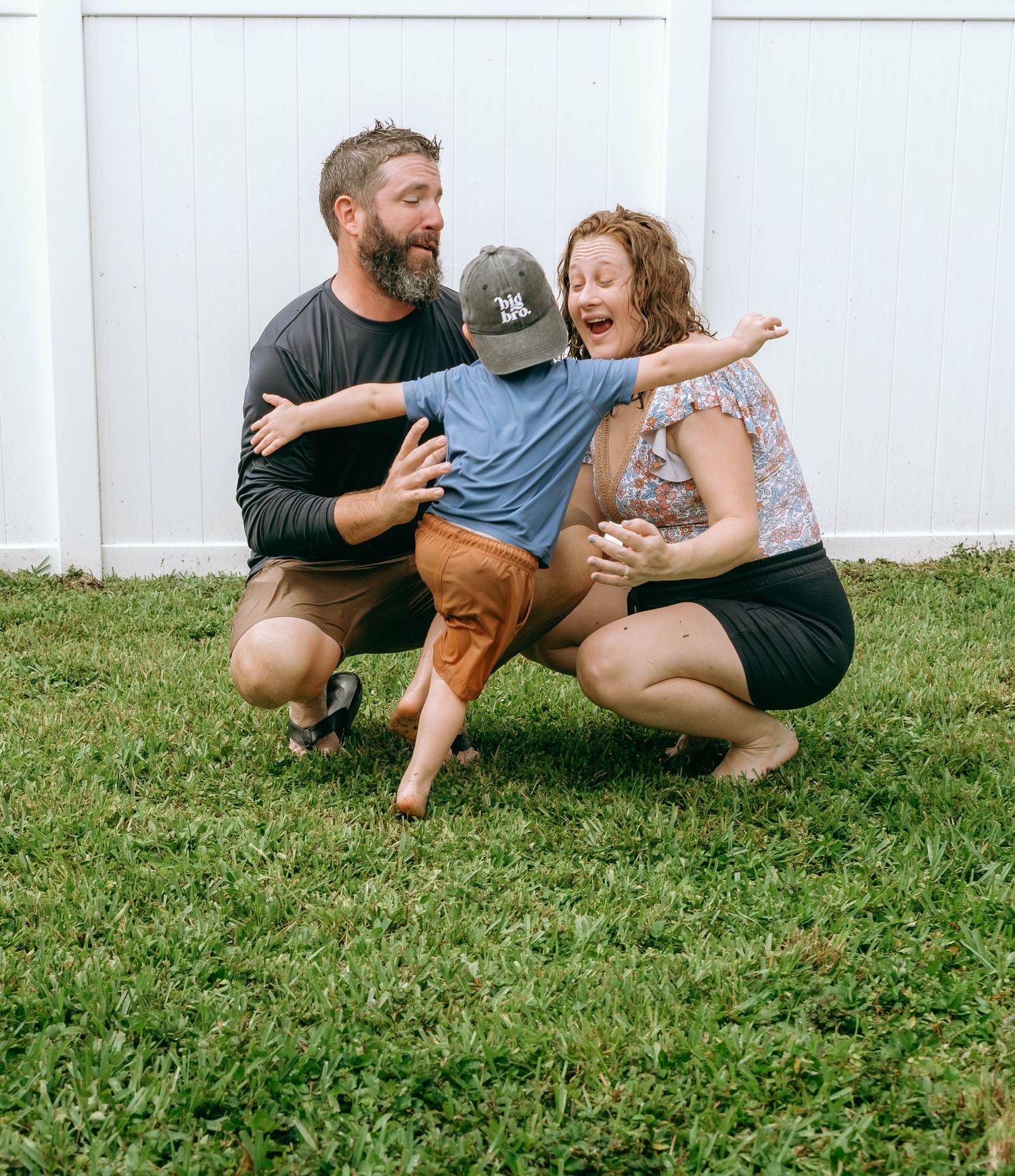 A joyful family of three laughs together on green grass, with parents crouching beside their young son wearing a cap.