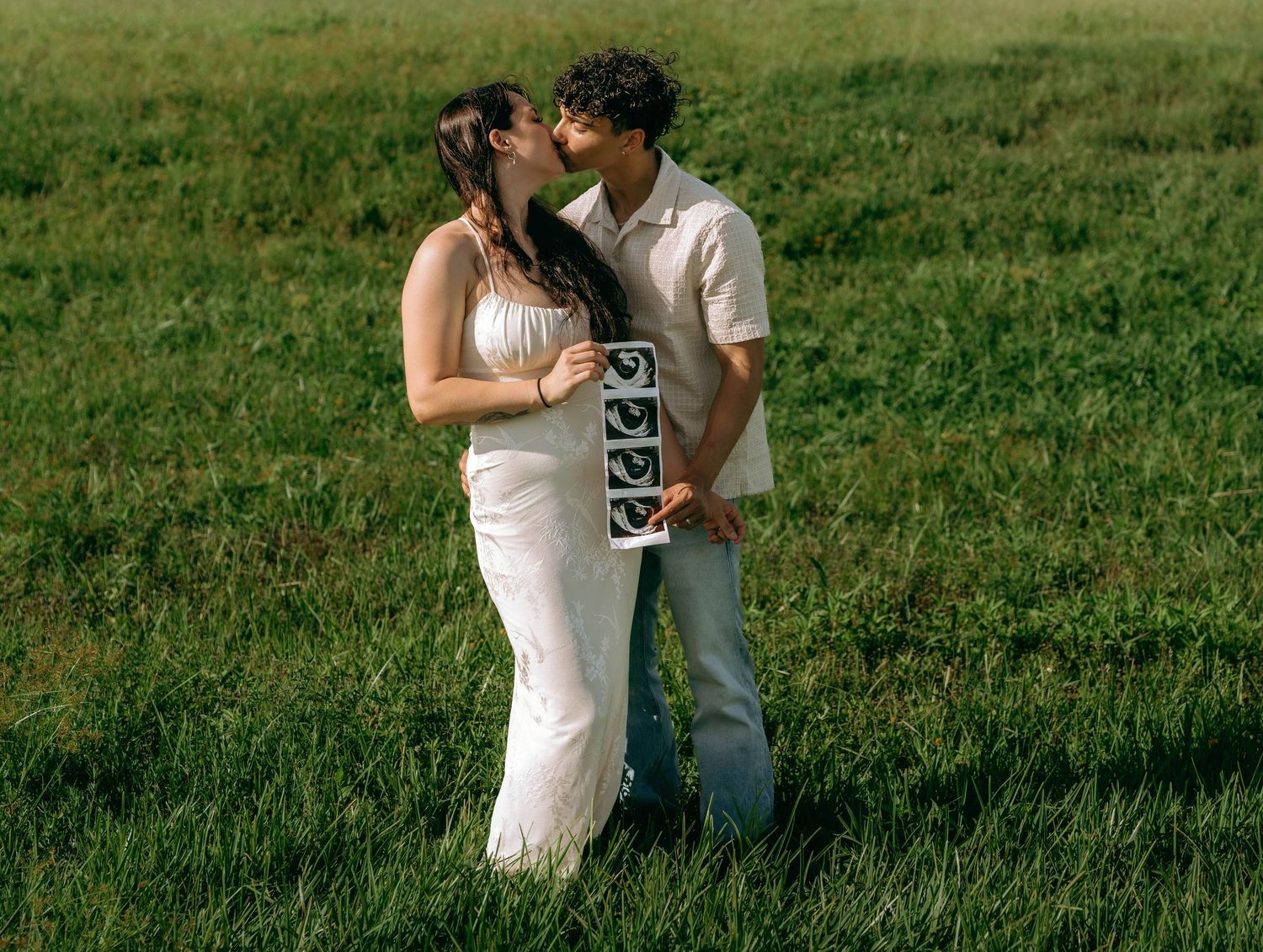 A couple kisses in a green field, the man holding ultrasound photos, wearing white outfits in a pregnancy announcement.