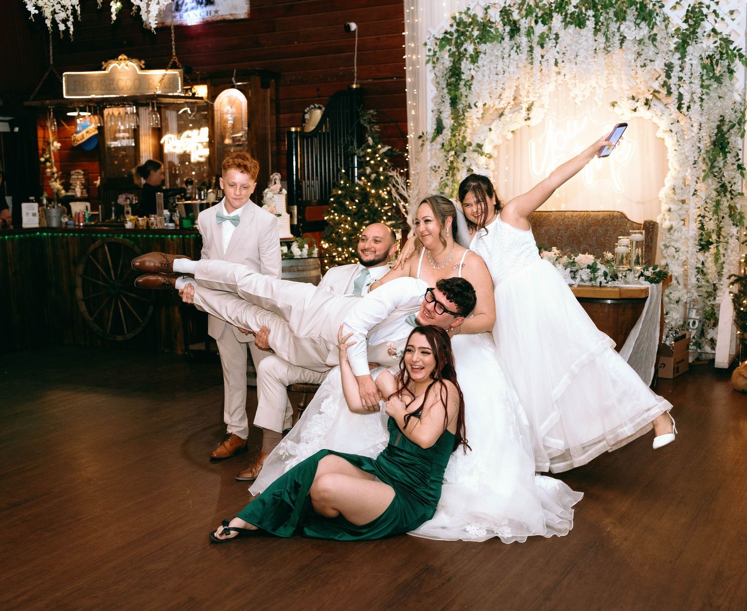 Wedding party members perform playful dance moves on a wooden dance floor during reception celebration.