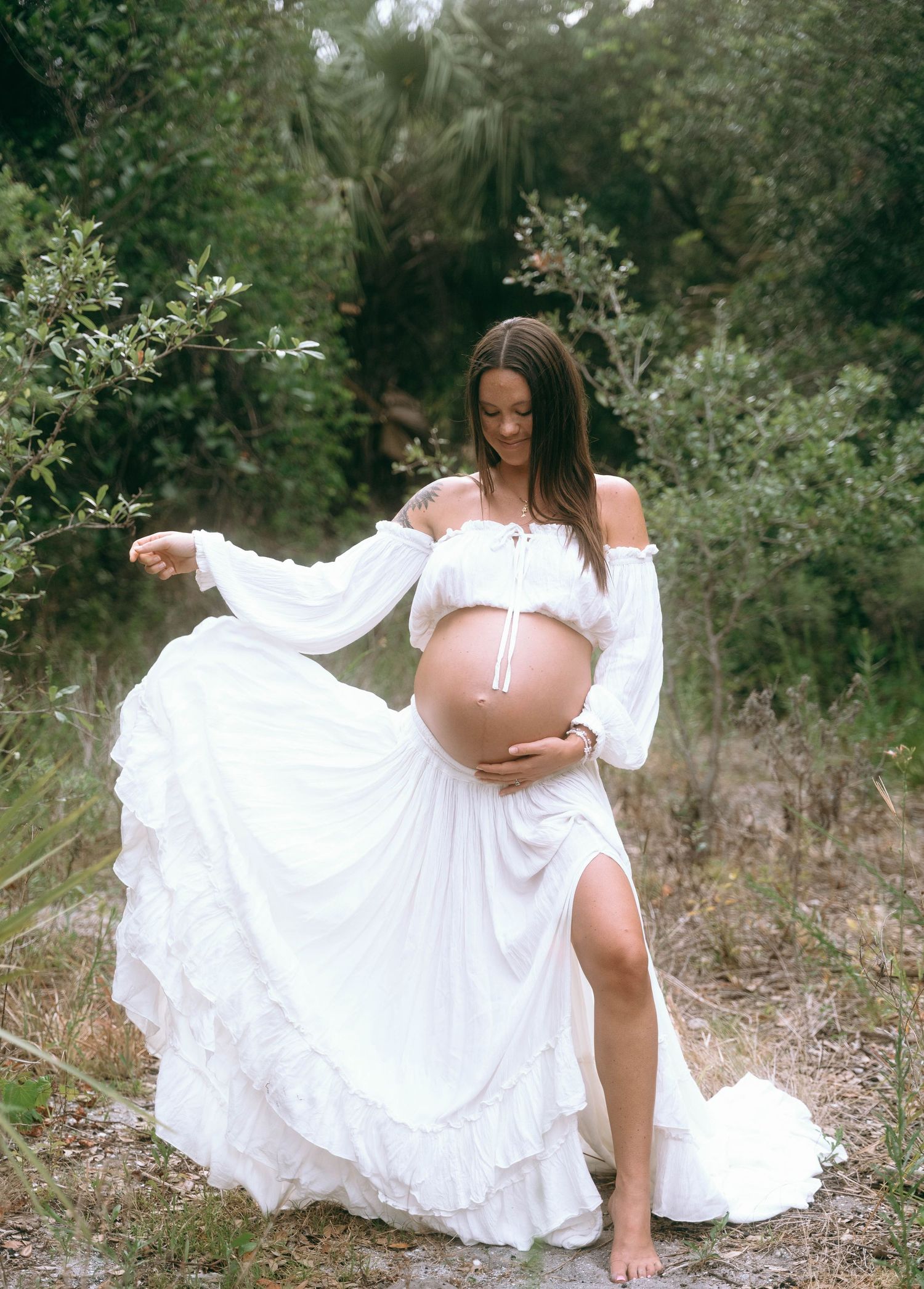 Ethereal maternity portrait in a flowing white dress outdoors among natural scenery.