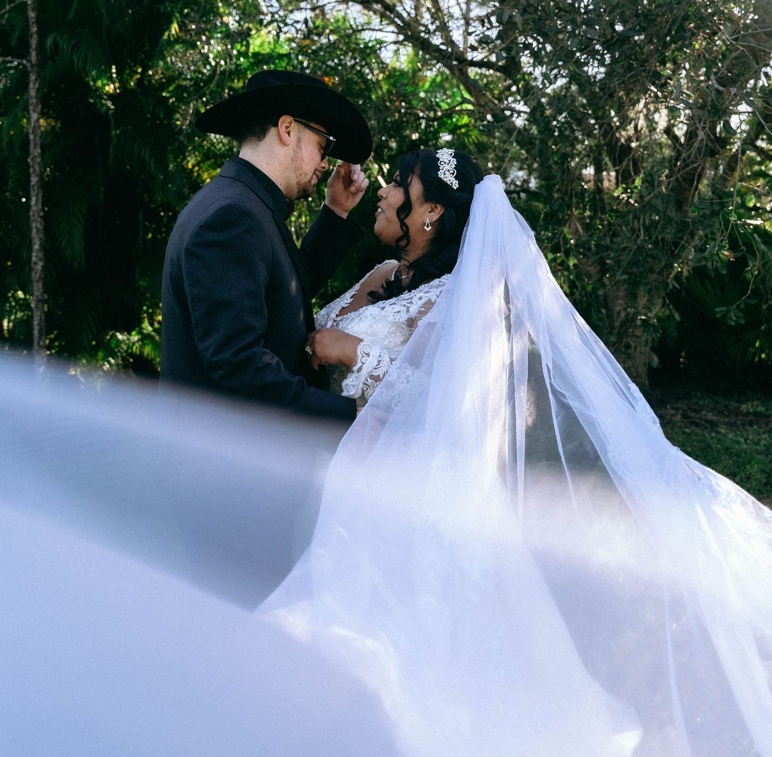 An outdoor wedding scene with flowing white veil in the foreground against a lush garden backdrop.