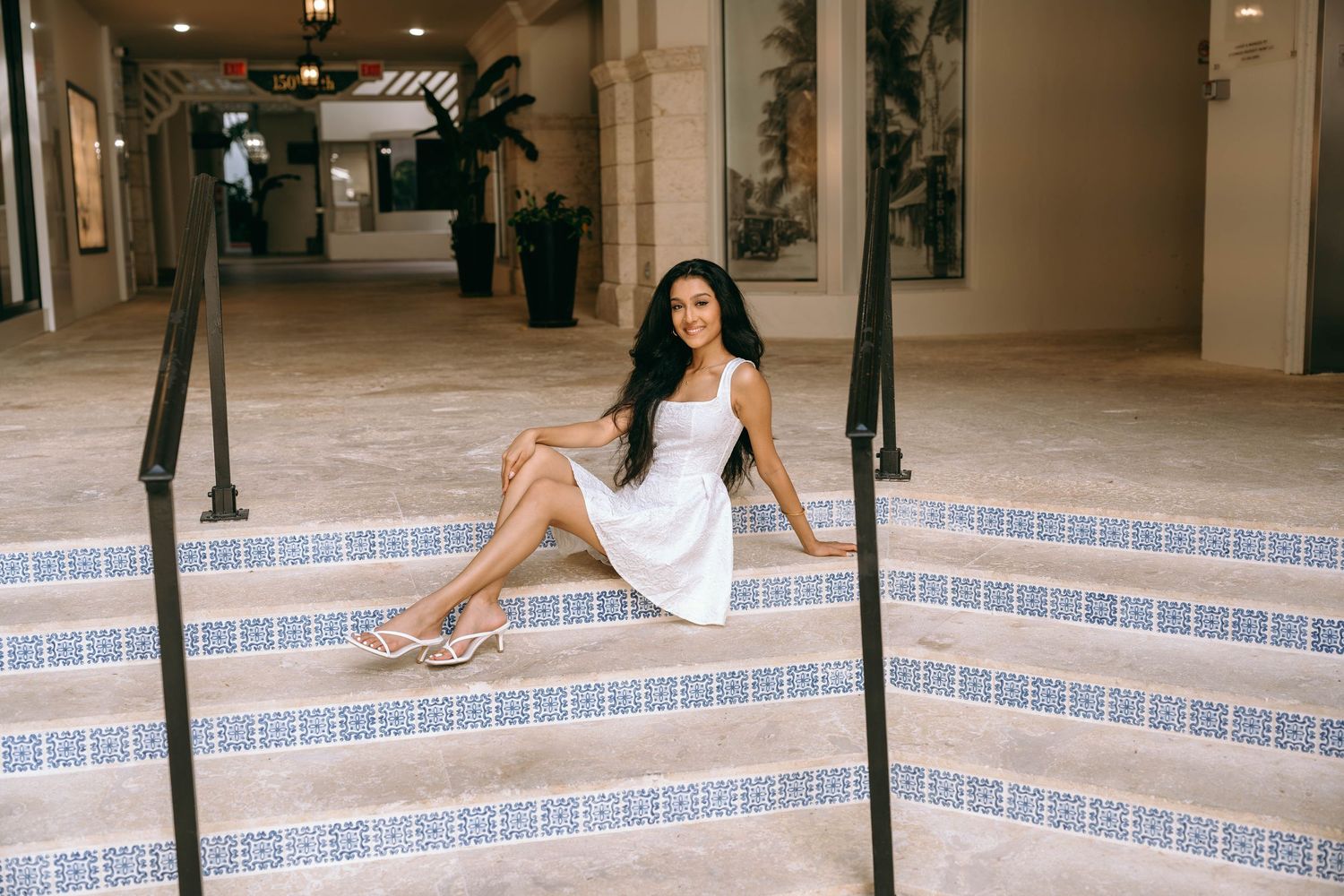 Young woman in white dress sitting on decorative tiled steps in an outdoor corridor smiling on Worth Avenue Palm beach.