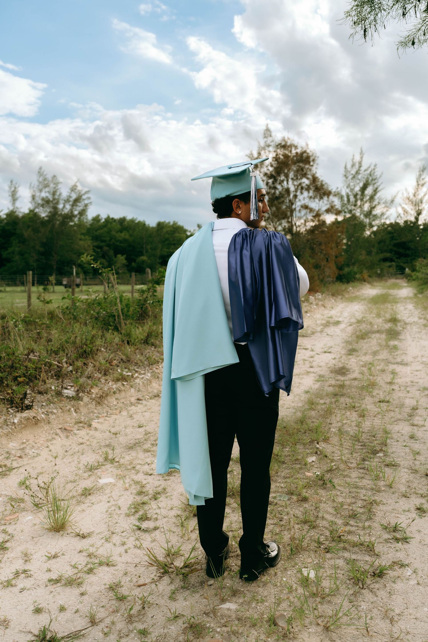 Graduate in light blue cap and gown holds navy robe while standing on sandy path surrounded by greenery. Senior Photos at Colt Farms