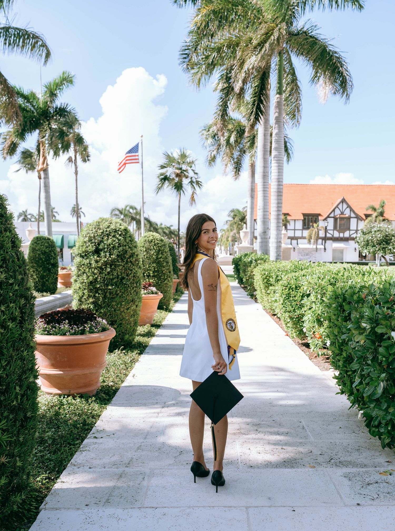 Smiling graduate in white dress with gold sash holding cap on palm-lined garden path on Worth Avenue Palm beach.