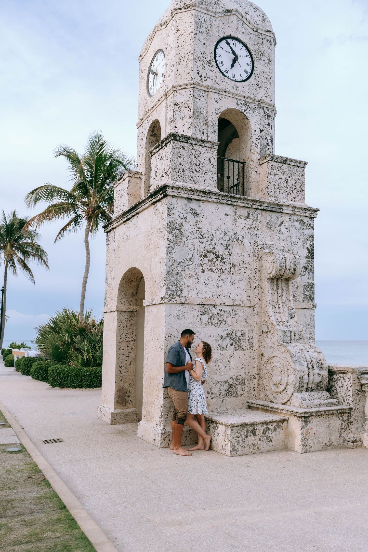 Loving couple embracing beside a historic stone clock tower near palm trees and the ocean at dusk on Worth Avenue Palm beach.