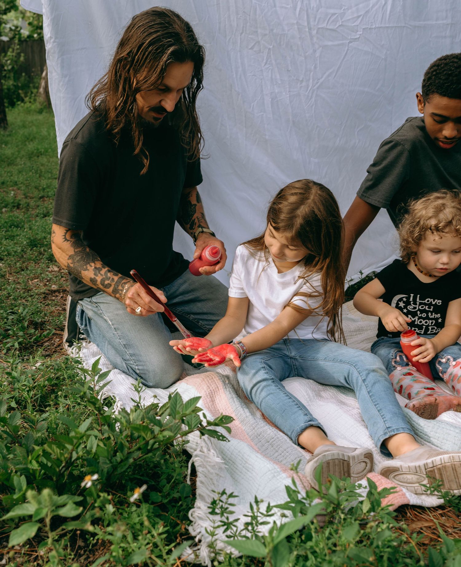 Tattooed man with long hair sits on a blanket outdoors with three children, sharing red fruit together.