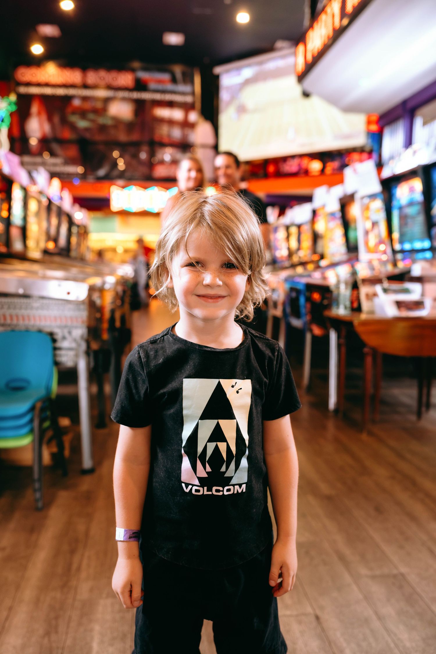 Young child smiling in a Volcom t-shirt inside a colorful arcade game room with adults in background.