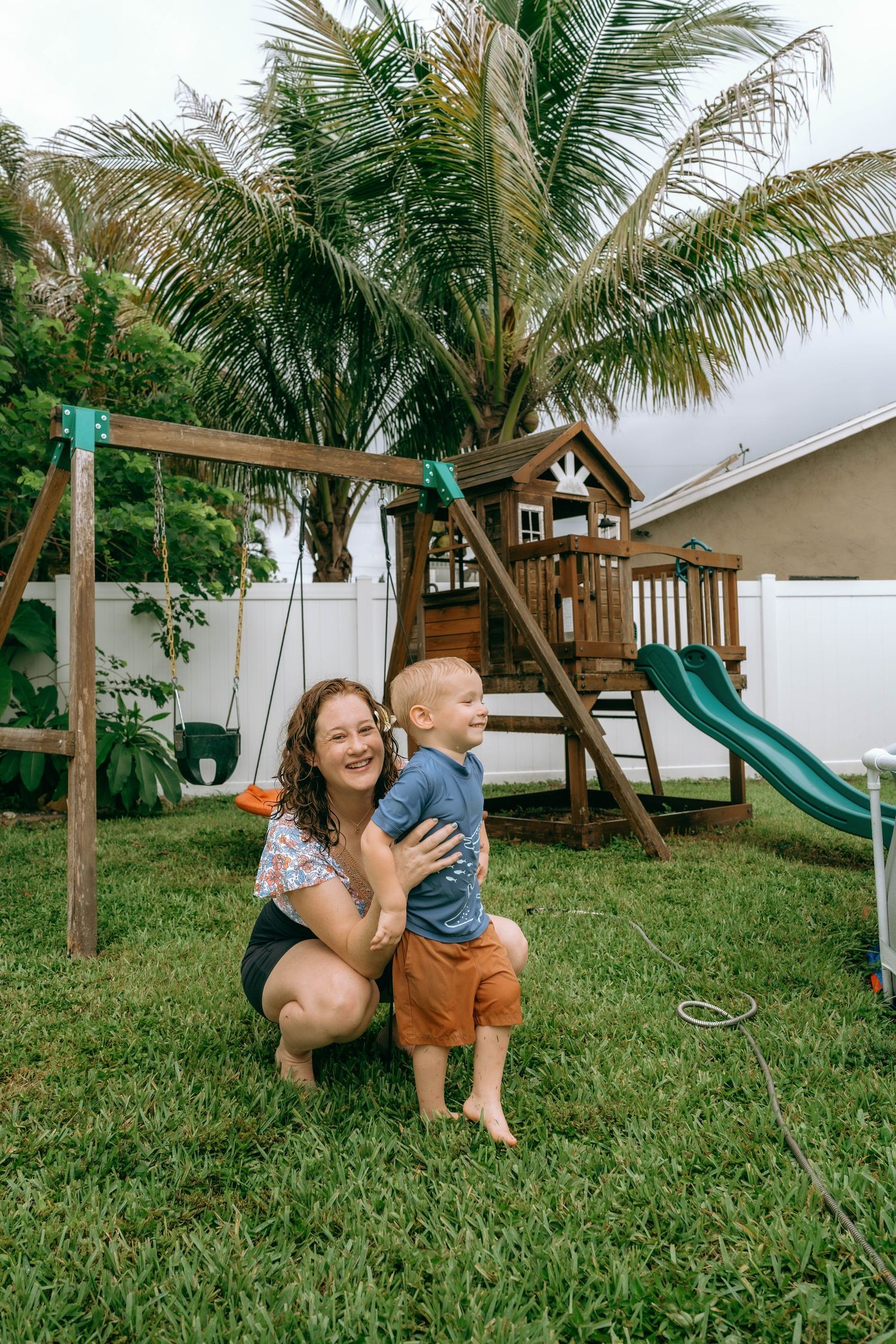 A smiling mother crouches with her toddler son in a backyard with a wooden playset and palm tree.