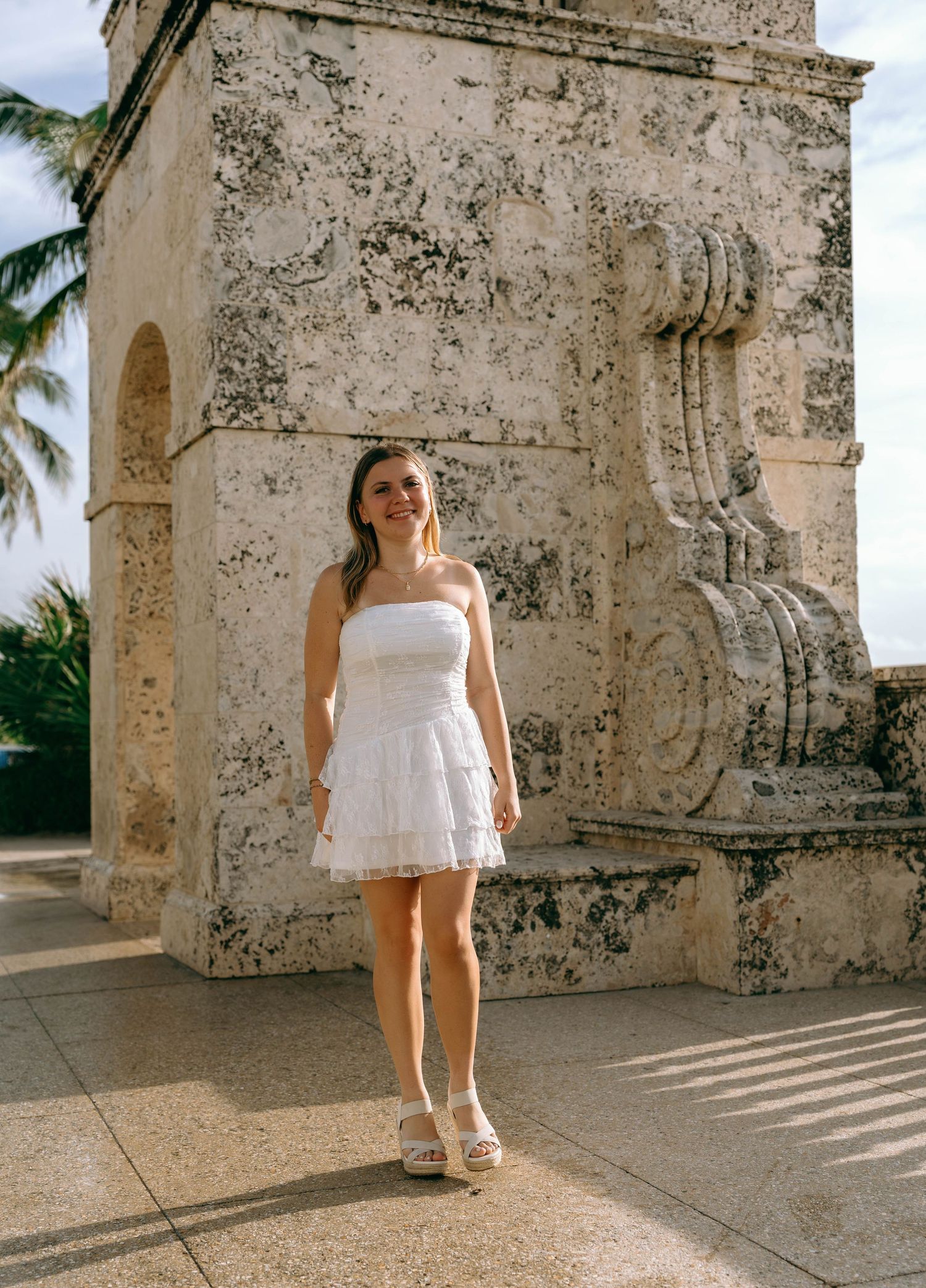 Young woman in white strapless mini dress and espadrilles posing by ornate stone monument outdoors.