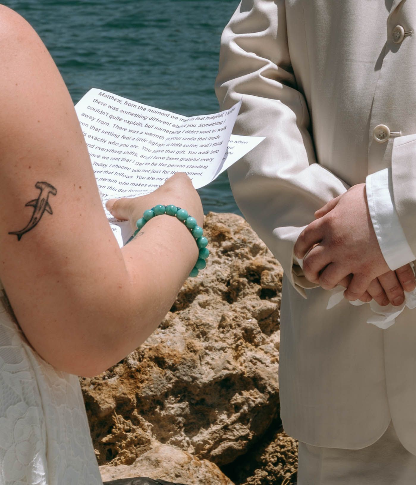 Bride with anchor tattoo and turquoise bracelet reads wedding vows by rocky ocean shore during outdoor ceremony.
