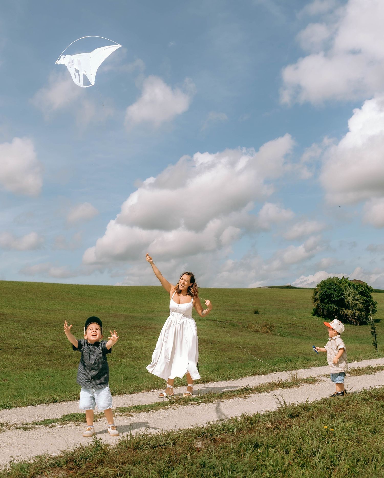 A woman in a white dress flies a kite with two young children on a sunny green hillside path.