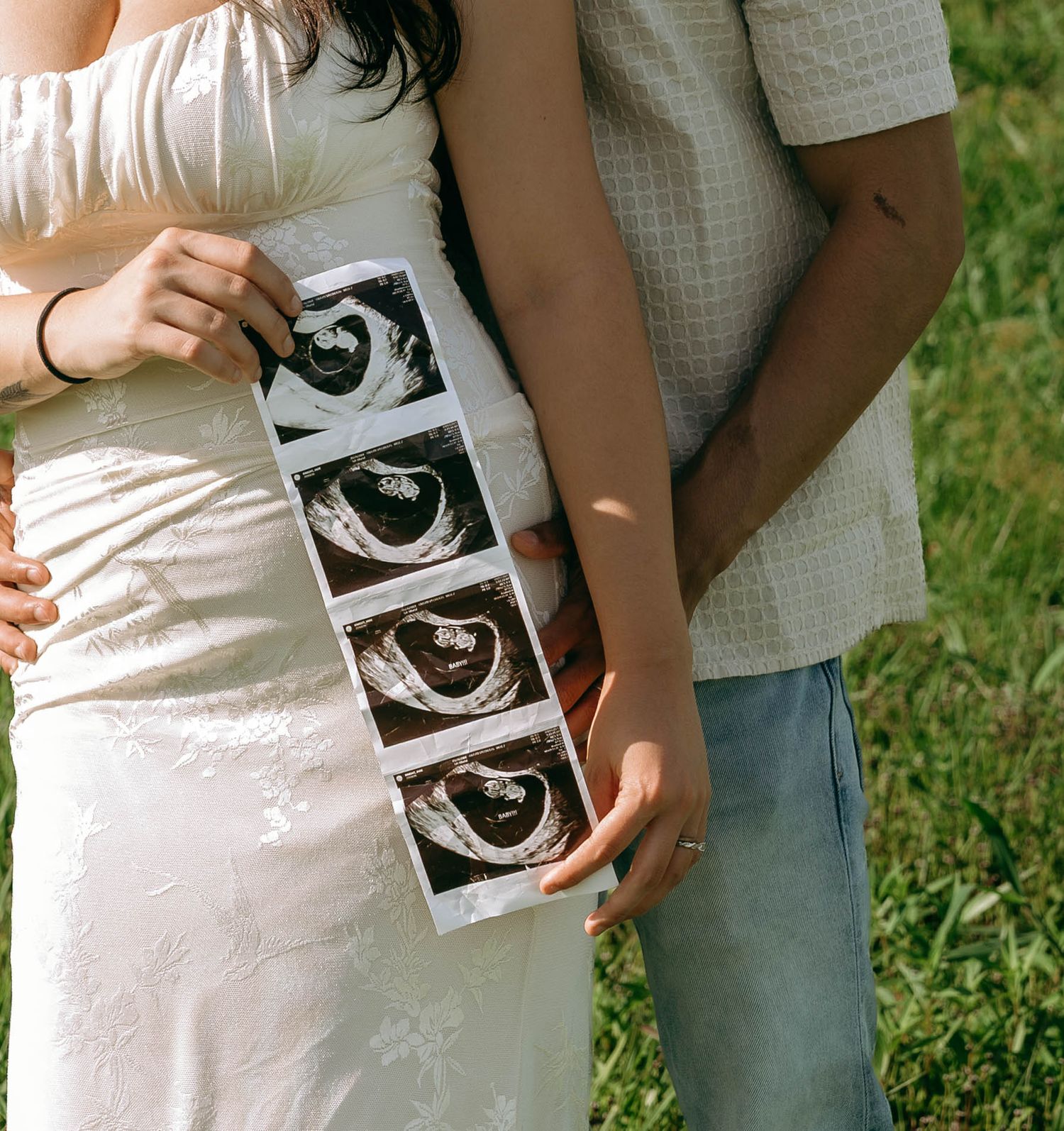 Pregnant couple holding ultrasound sonogram photos outdoors, woman in white floral dress standing on green grass.