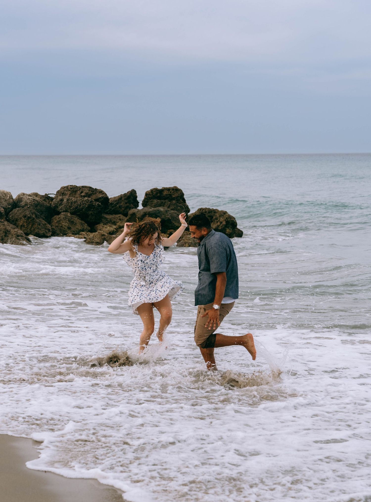 Happy couple splashing and running through ocean waves on a rocky beach under a cloudy sky on Worth Avenue Palm beach.
