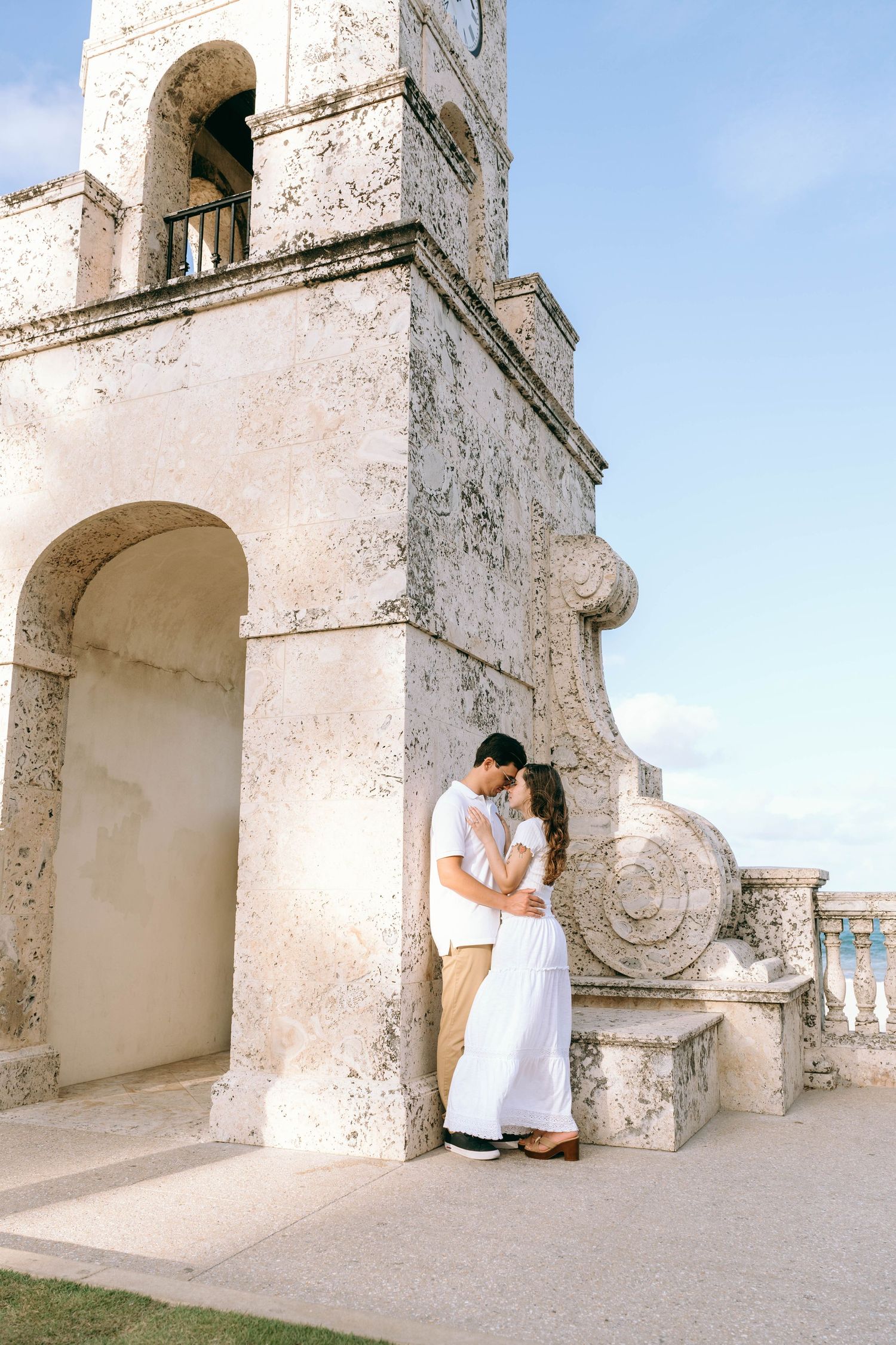Couple embracing romantically beside a historic stone clock tower under a clear blue sky. Engagement photos on Worth Avenue in Palm Beach