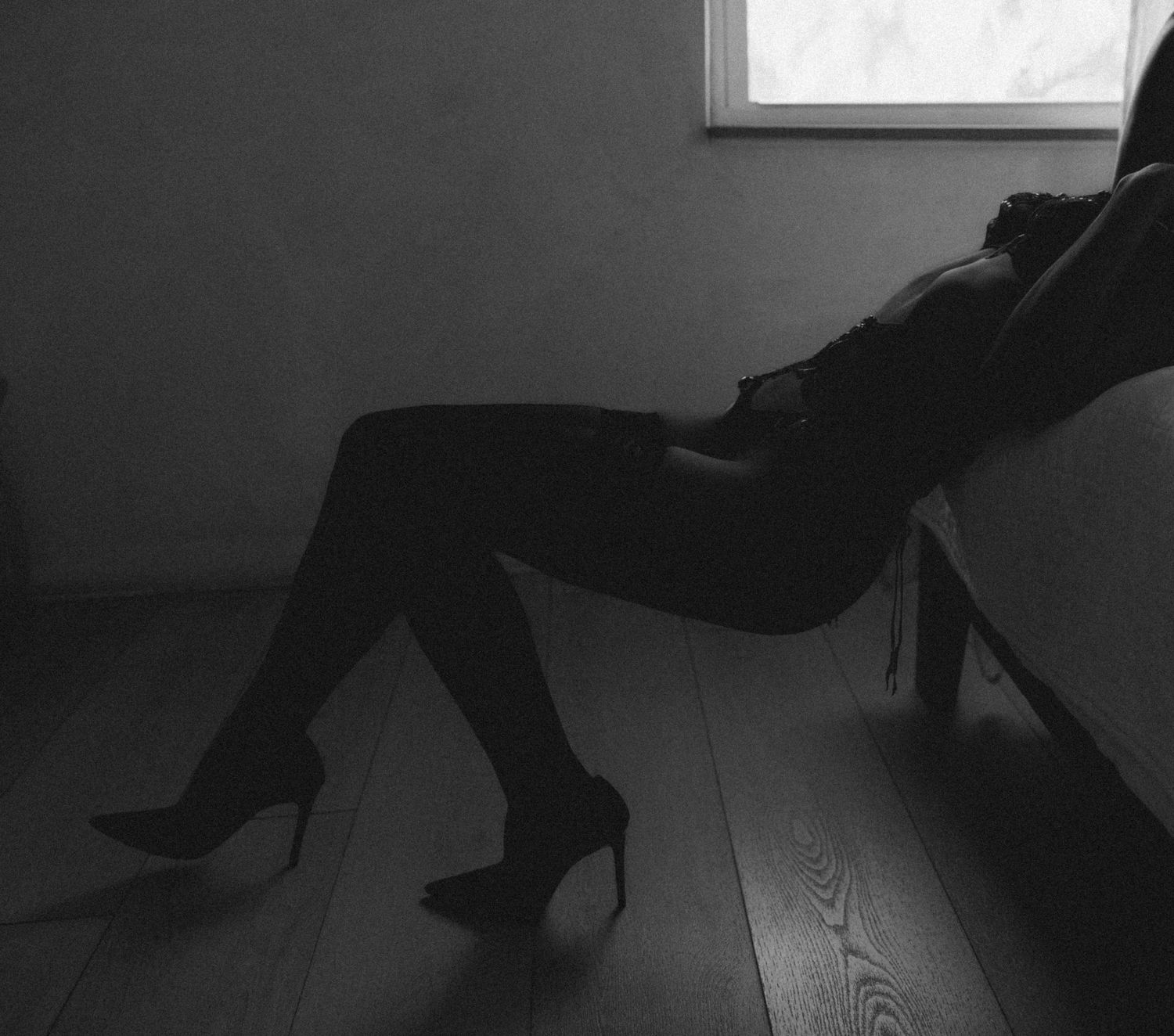 Black and white photo of a woman in heels and dark clothing seated in a chair near a bright window.