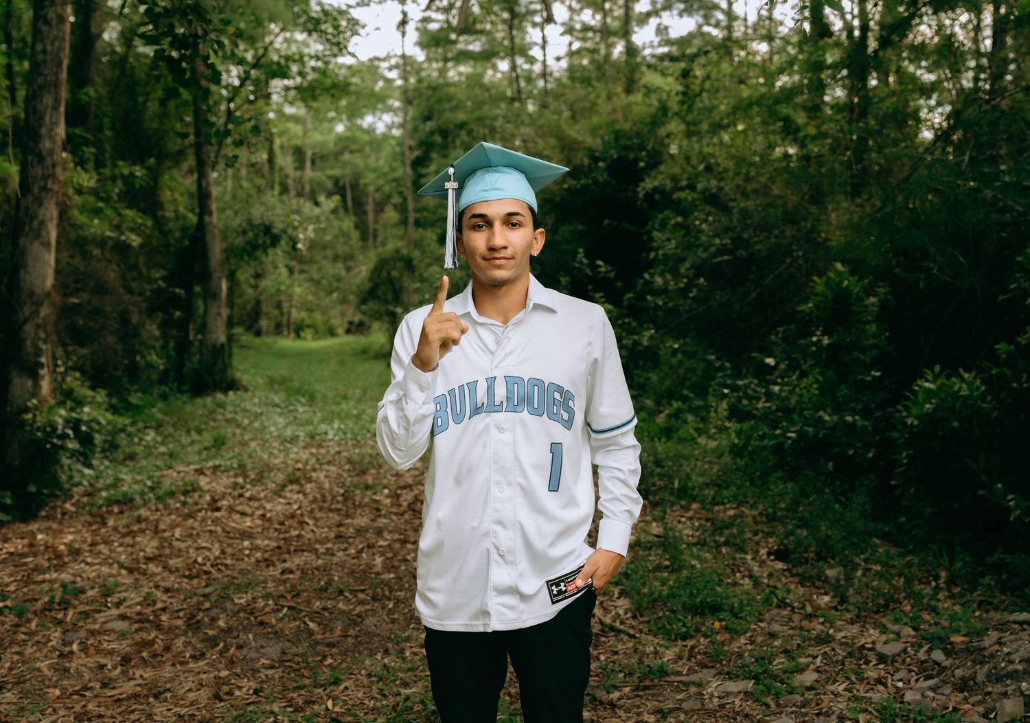 Young man in Bulldogs jersey and teal graduation cap posing outdoors in a forest setting. Senior Photos at Colt Farms in Boynton Beach, FL