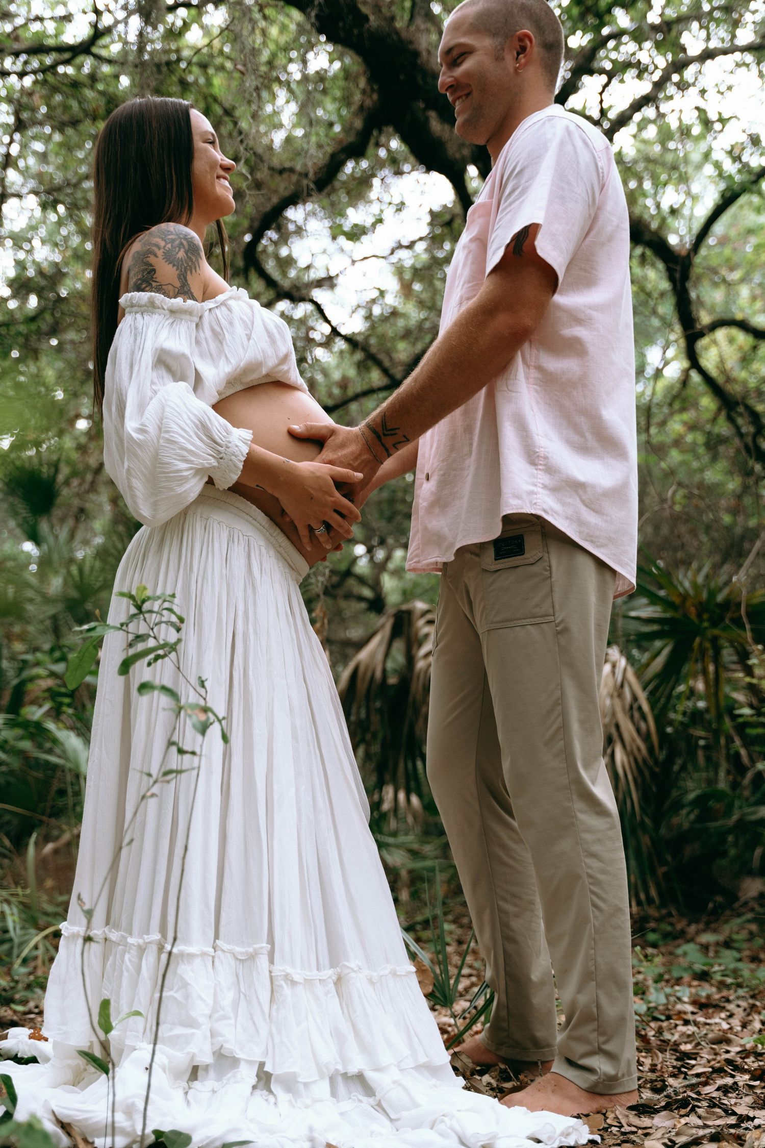 Couple in white and tan outfits share tender moments during forest photoshoot.