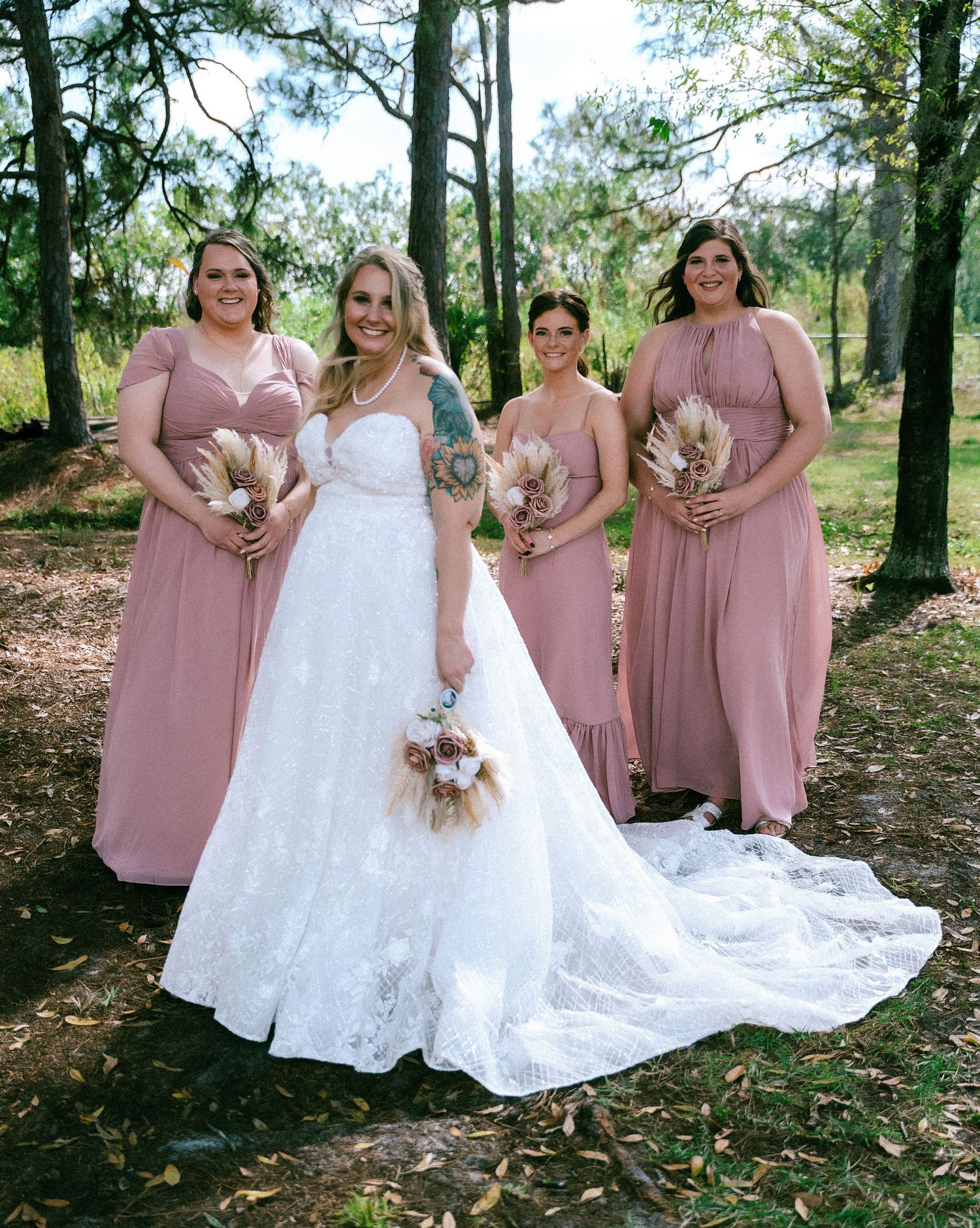 Bride in white lace gown poses with three bridesmaids in dusty rose dresses outdoors among trees.