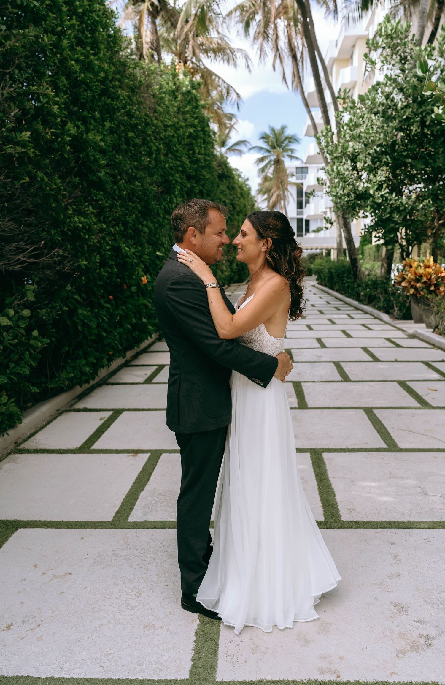 Bride and groom embrace on elegant stone pathway surrounded by lush tropical greenery at outdoor wedding venue.