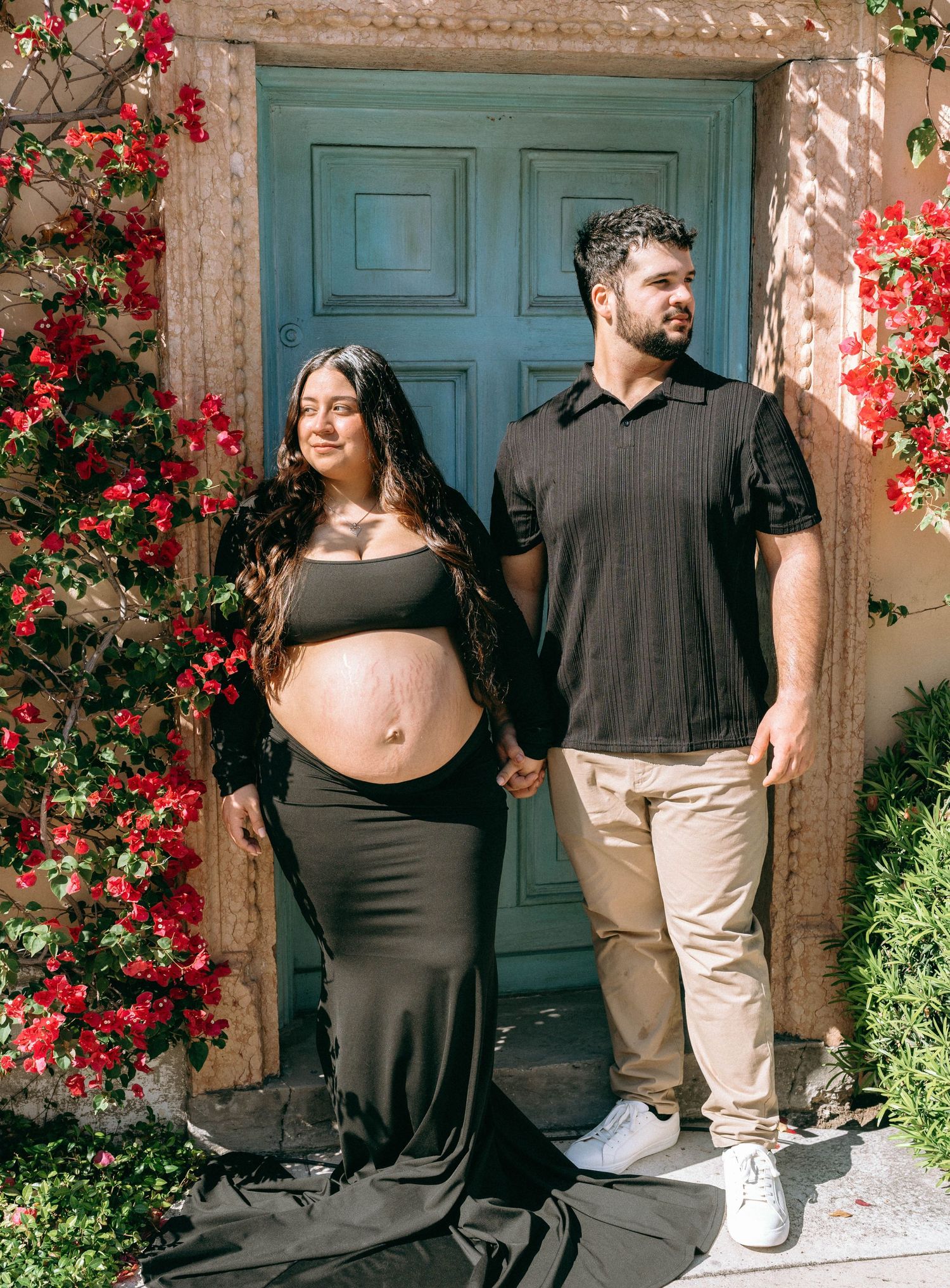 Pregnant couple holding hands before teal door with red bougainvillea, woman in black maternity gown. Worth Avenue, Palm Beach, FL