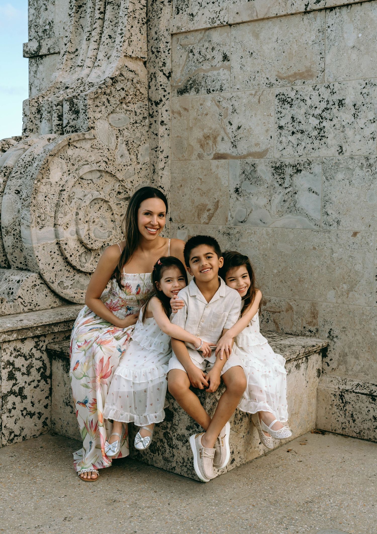 A smiling mother in a floral dress sits with her three children on stone steps near ornate carved architecture. Worth Avenue, Palm Beach, FL