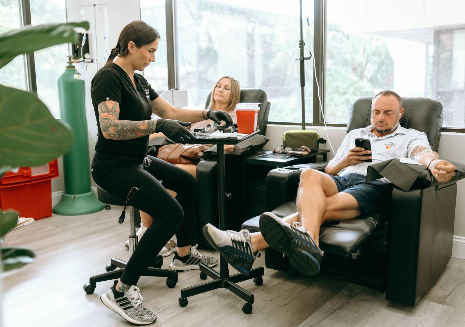 Tattoo artist working on client in modern studio while another person relaxes in recliner chair using smartphone.