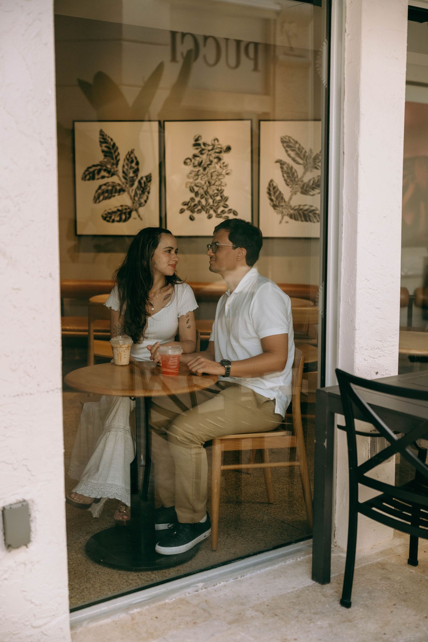 Couple sharing drinks at a cozy cafe table, gazing at each other with botanical art prints on the wall behind them on Worth Avenue Palm bea.
