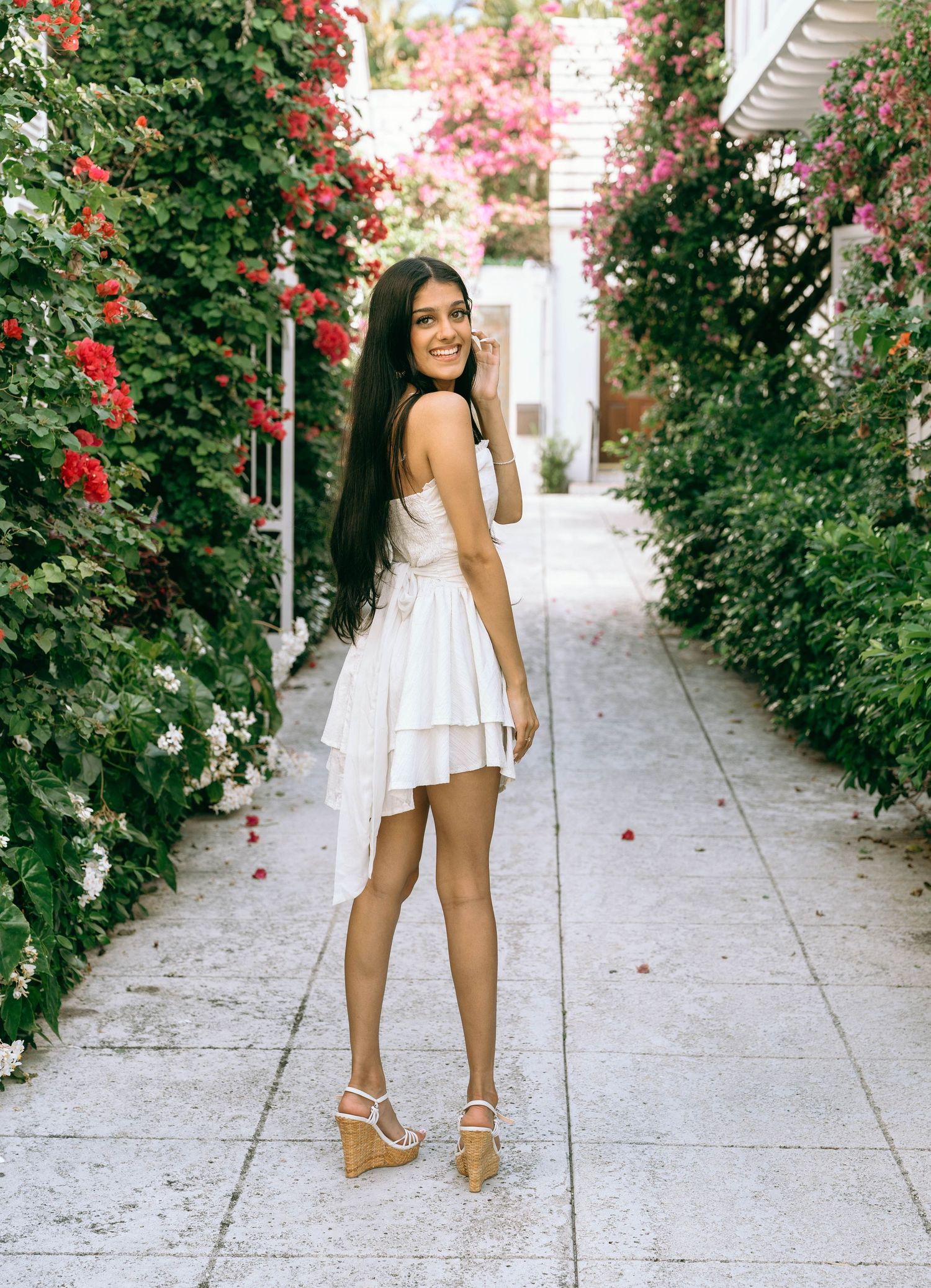 Young woman in white ruffled mini dress and wedge sandals posing in a flower-lined garden pathway on Worth Avenue Palm beach.