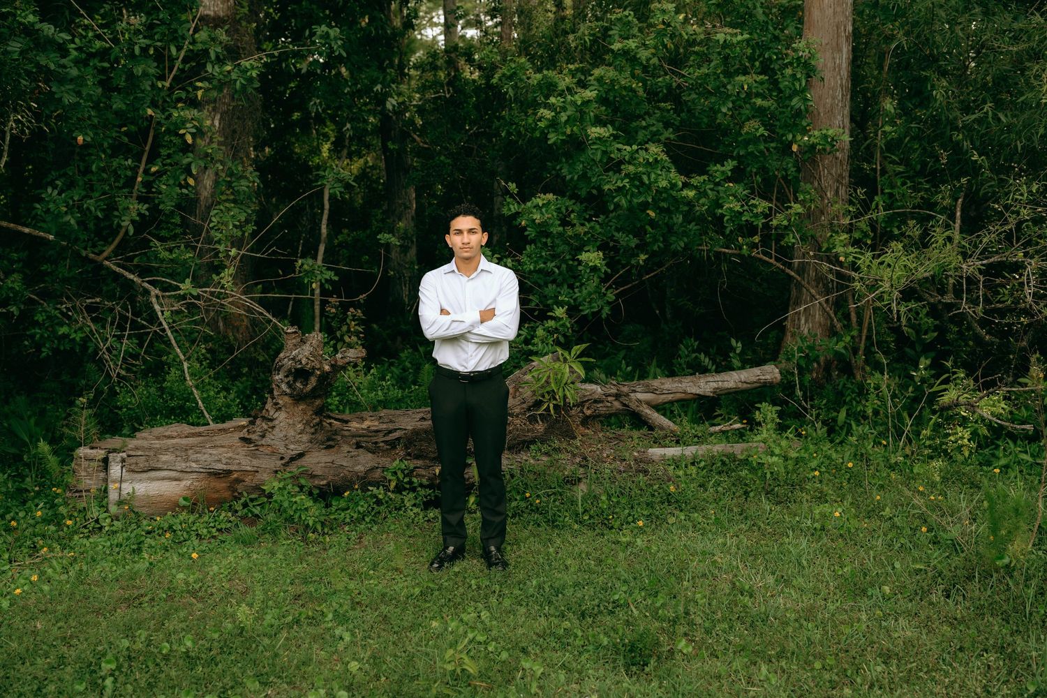 A young man in a white shirt and black pants stands with arms crossed in a lush green forest clearing. Senior Photos at Colt Farms