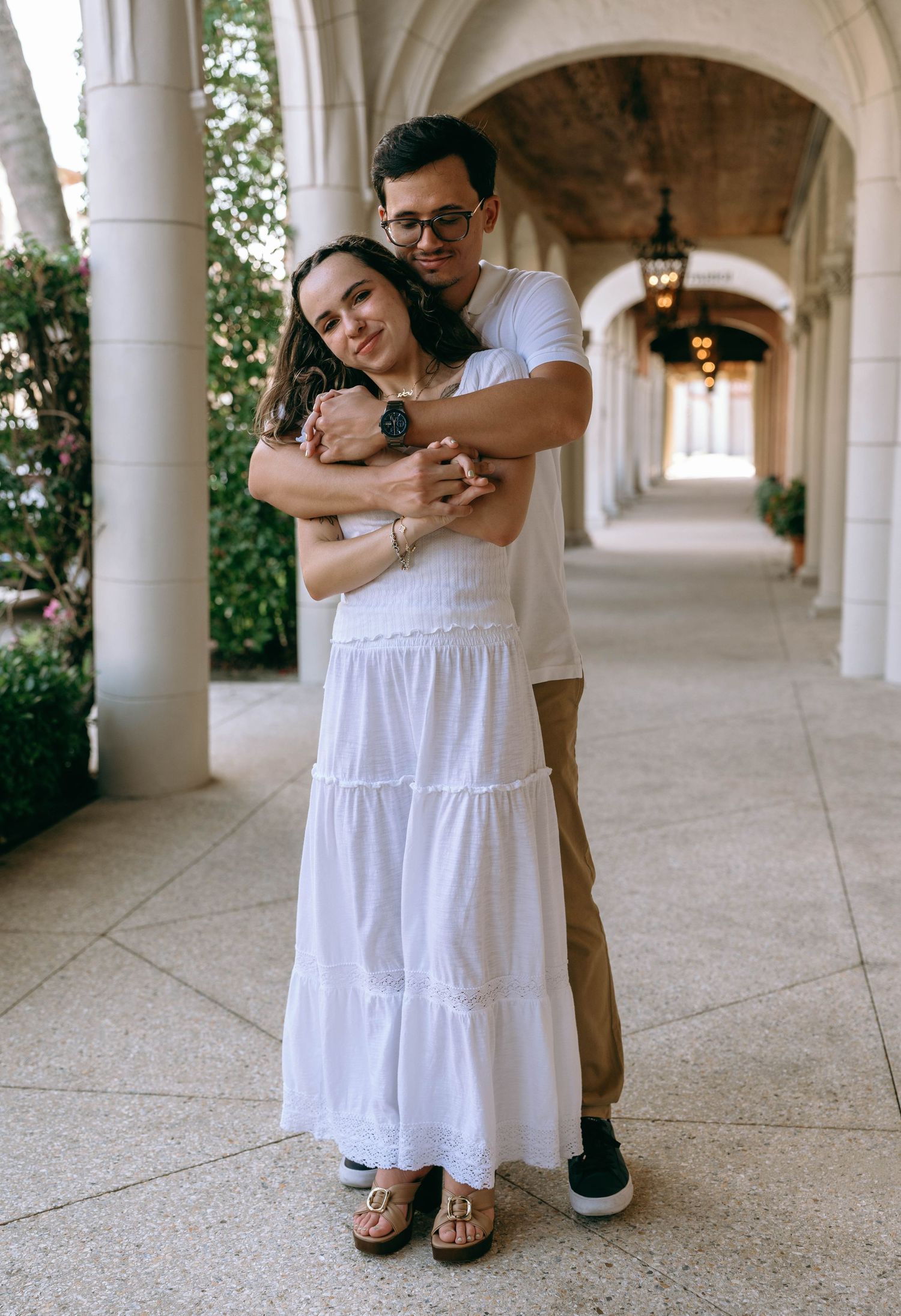 A couple in white outfits embrace lovingly in an elegant arched corridor with warm lighting. Engagement photos on Worth Avenue in Palm Beach
