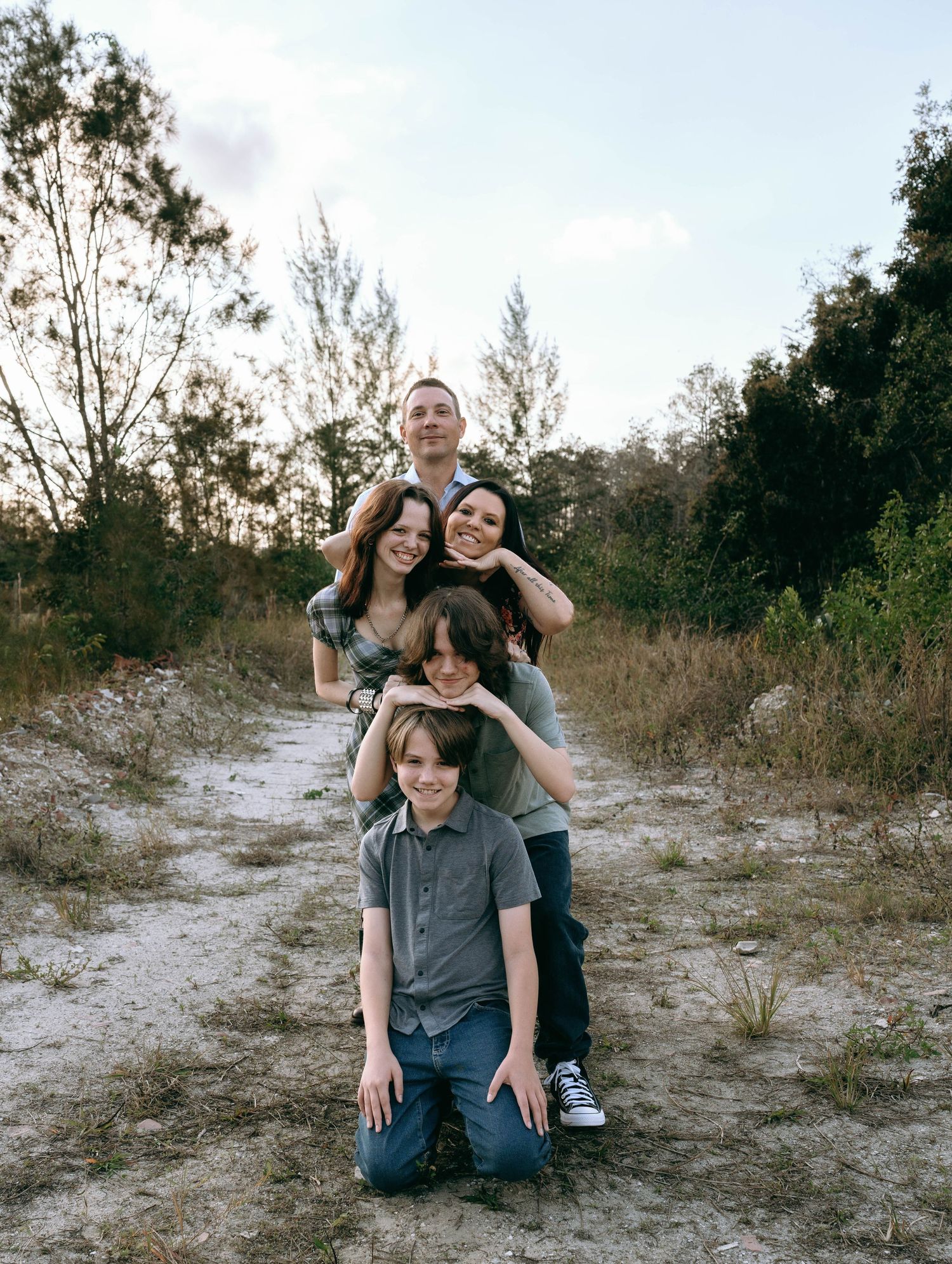 A joyful family of five poses playfully on a sandy trail surrounded by trees and golden grass outdoors at Colt Farms in Boynton Beach FL..