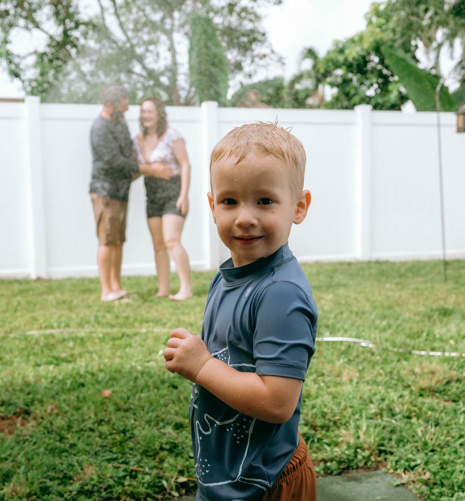 A smiling toddler boy in a blue shirt stands in a backyard while two adults play in a sprinkler behind him.